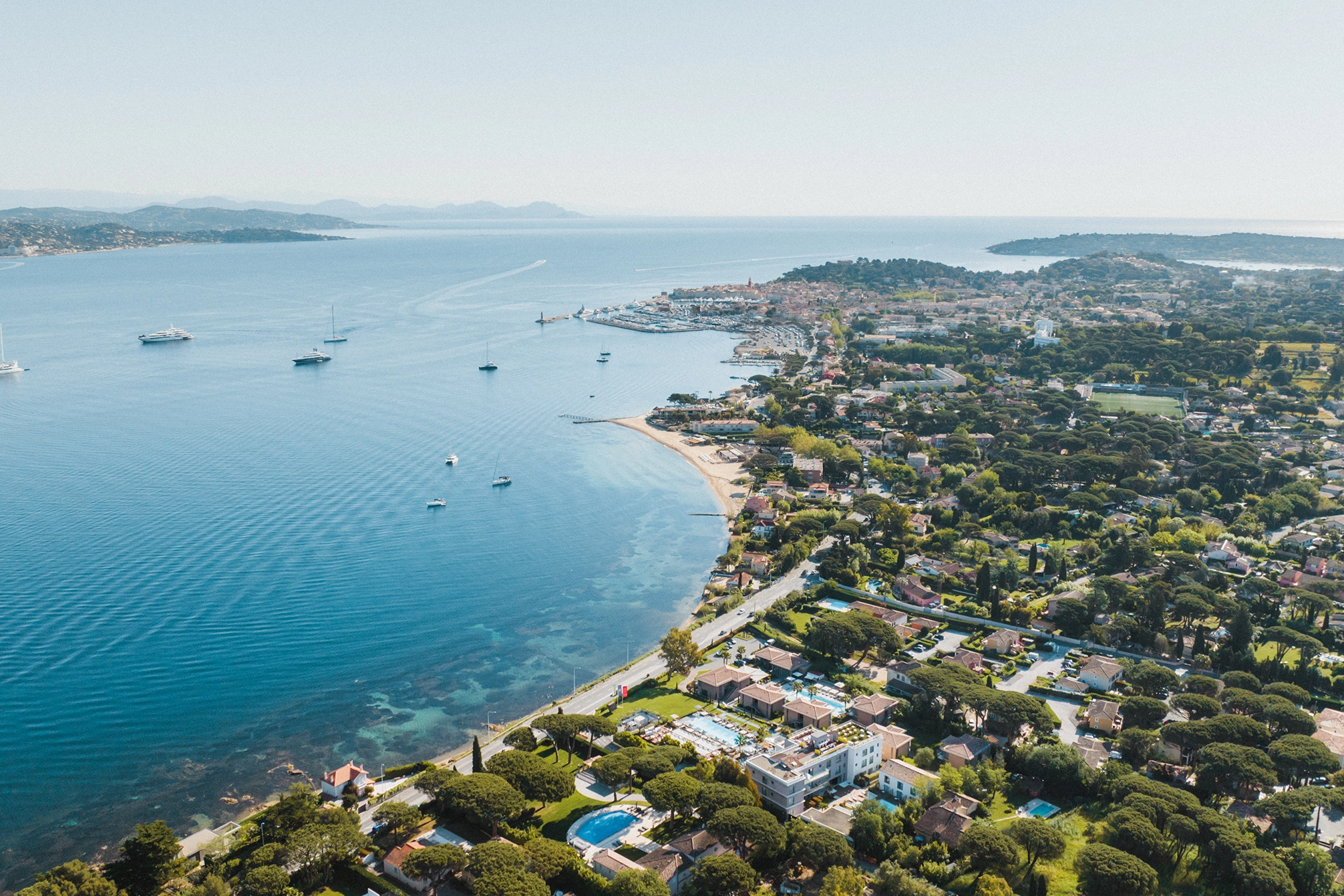Vue aérienne de la côte avec de nombreuses maisons, une plage, plusieurs bateaux ancrés dans une mer bleue calme, et une ville au loin.