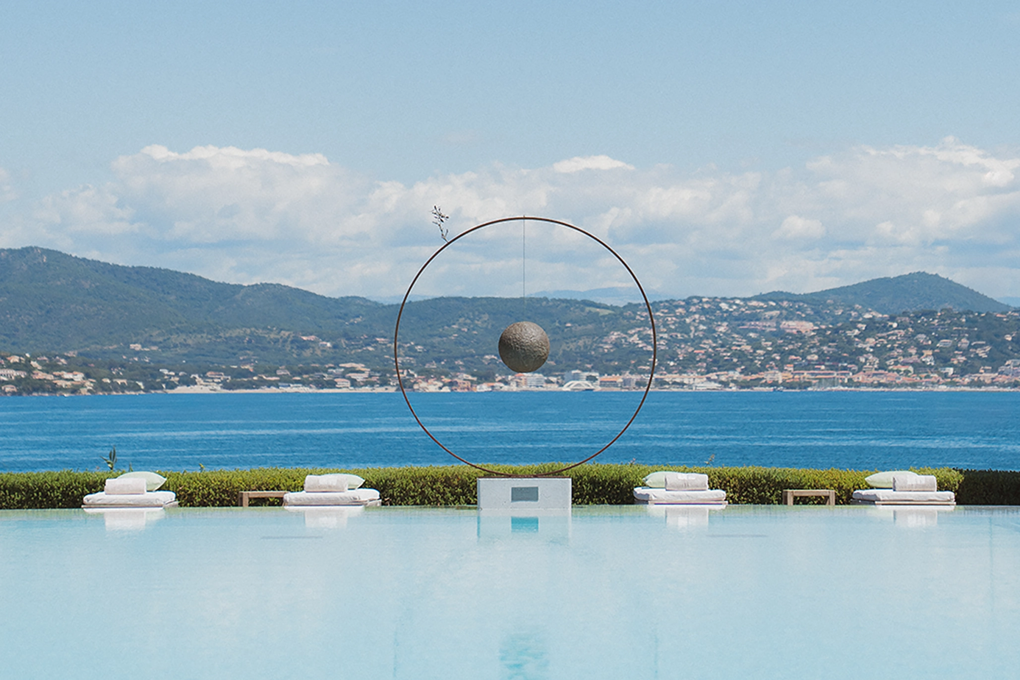 Piscine à débordement donnant sur la mer avec un cercle métallique et une sphère suspendue au centre, entourée de quatre chaises longues avec des serviettes.