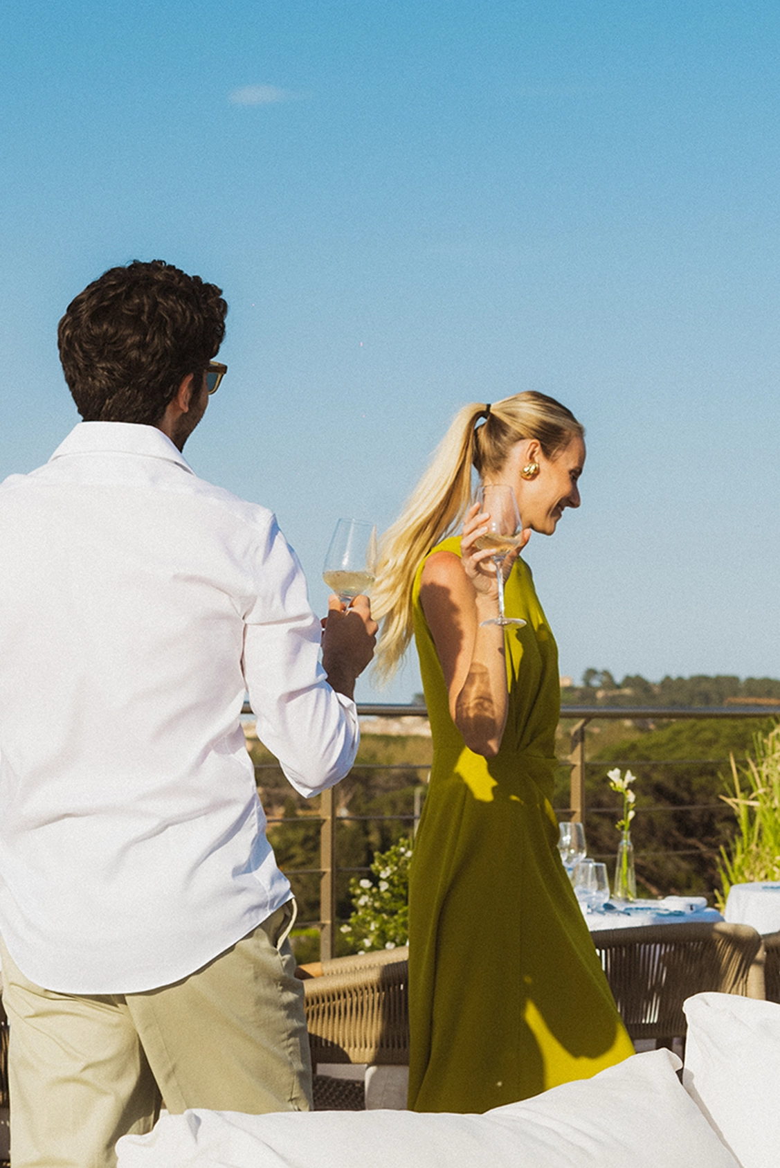 Un homme en chemise blanche et une femme en robe jaune tenant des verres de vin blanc sur une terrasse ensoleillée avec vue sur la nature.