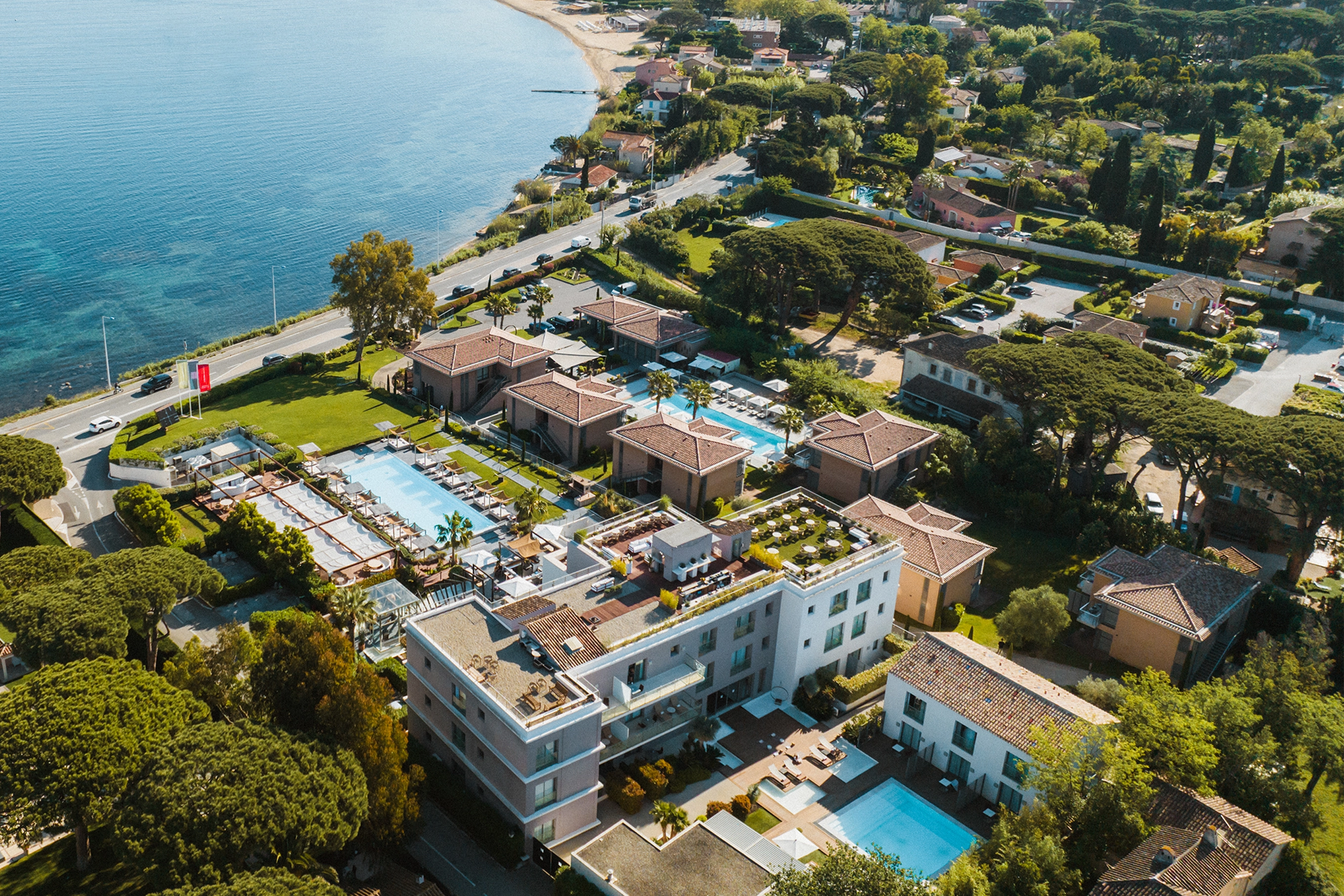 Vue aérienne d'un complexe hôtelier avec plusieurs piscines situé au bord d'une mer bleue, entouré de bâtiments, d'arbres et d'une route.
