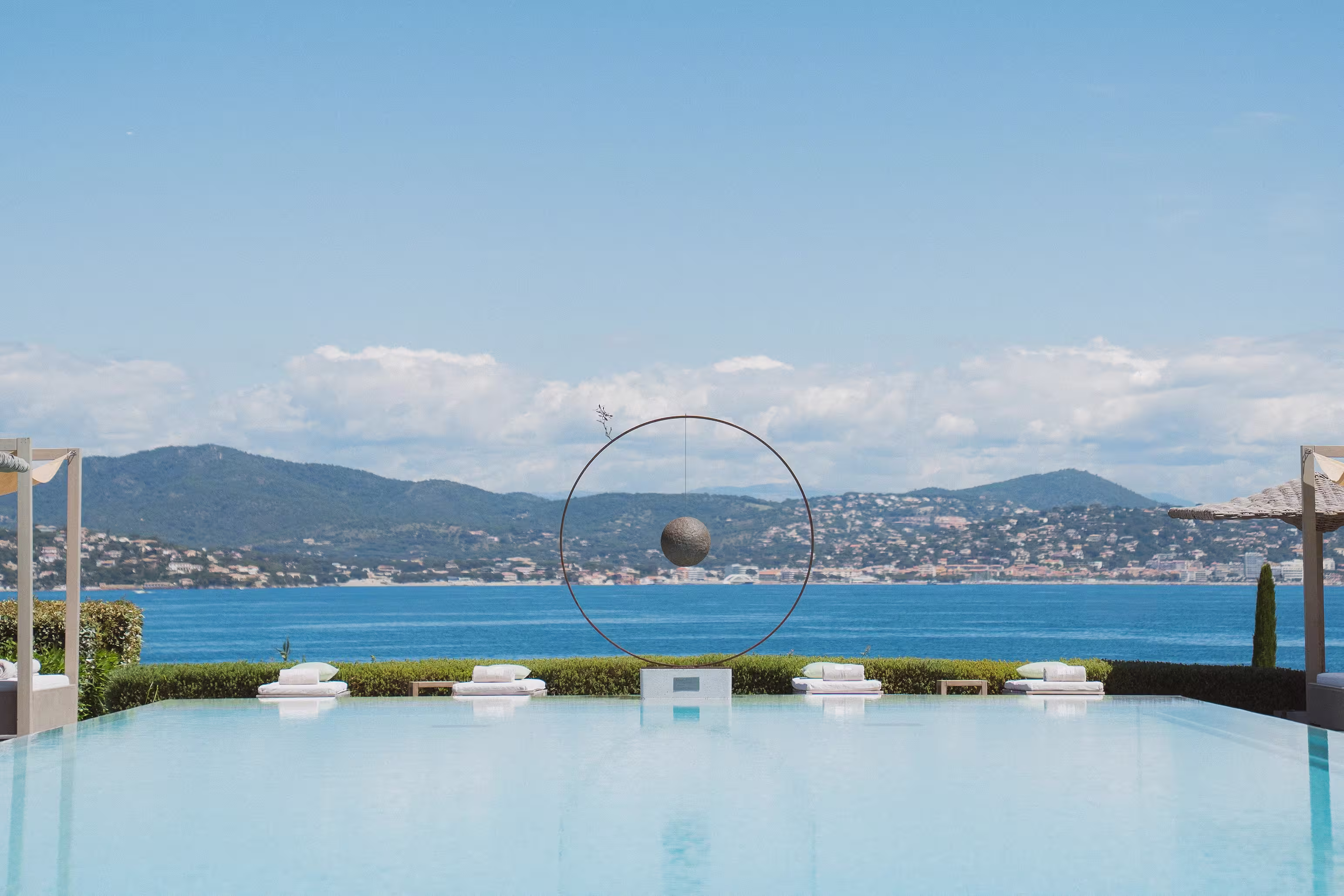Piscine à débordement avec quatre chaises longues et une sculpture ronde suspendue, vue sur la mer et les collines au loin.