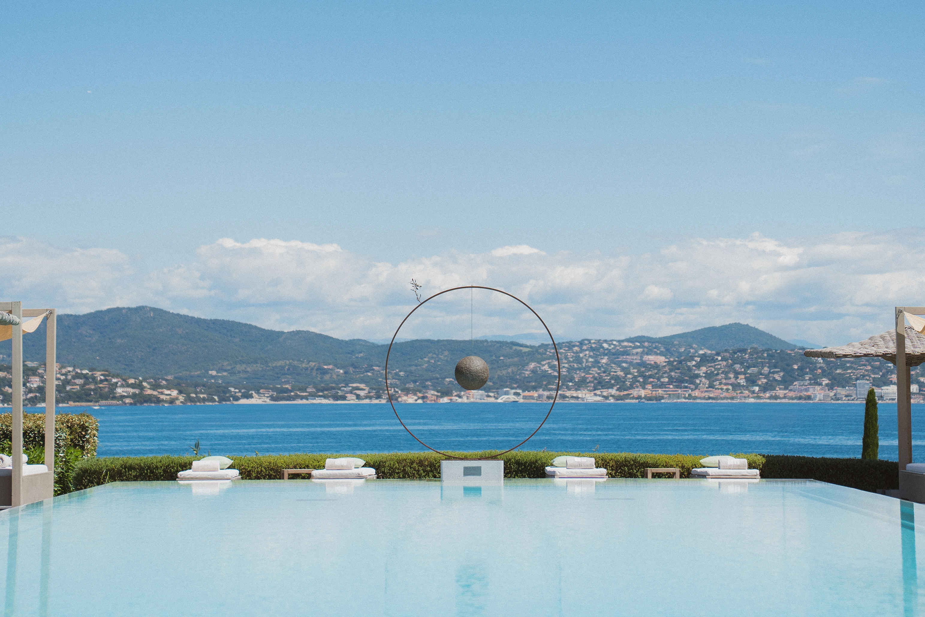 Piscine à débordement avec quatre chaises longues et une sculpture ronde suspendue, vue sur la mer et les collines au loin.