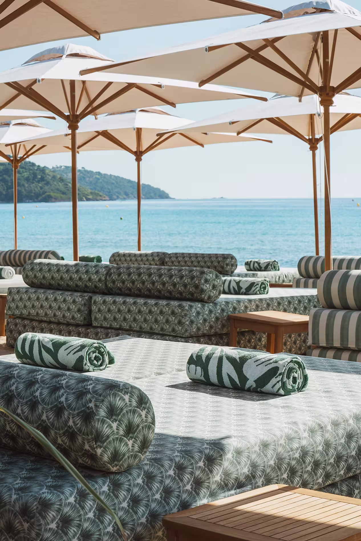Lits de plage avec coussins verts à motifs sous plusieurs parasols blancs face à la mer avec une colline en arrière-plan.