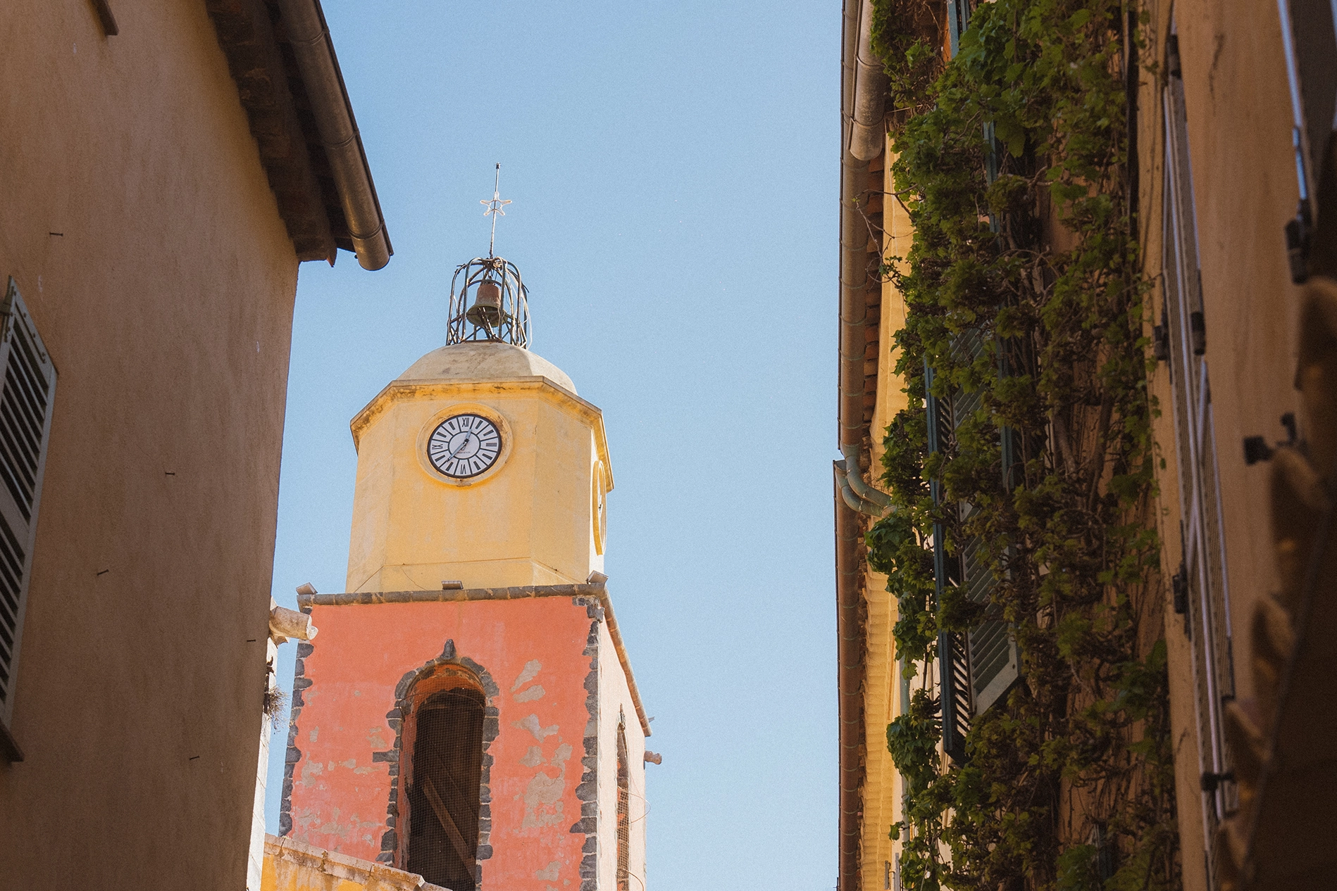 Tour d'horloge jaune et rose avec une cloche, entre deux bâtiments, sous un ciel bleu clair.
