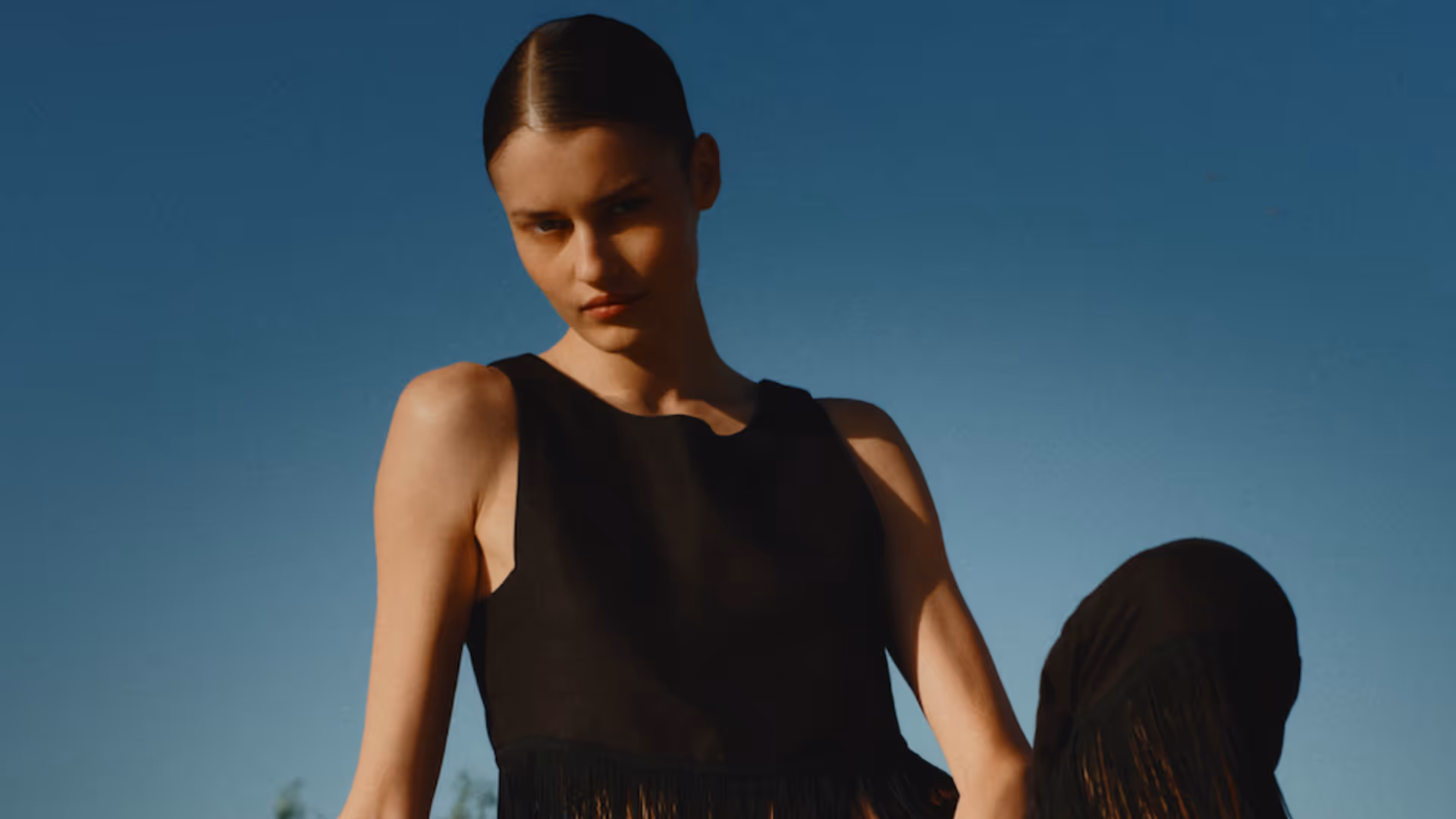 Jeune femme portant une robe noire sans manches contre un ciel bleu clair.