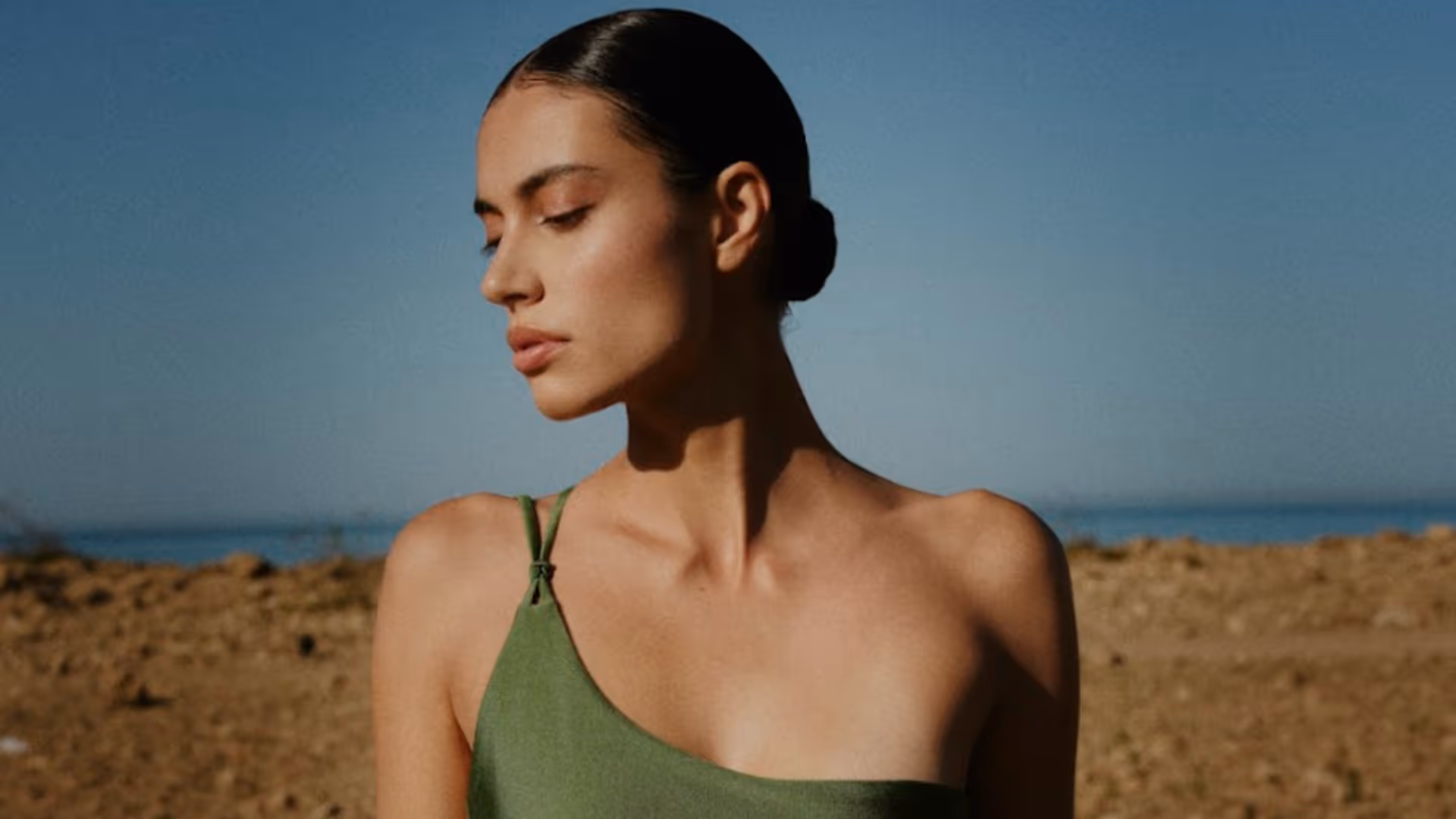 Femme aux cheveux noirs coiffés en chignon, portant une robe verte à bretelle, regardant sur le côté avec un ciel bleu en arrière-plan.