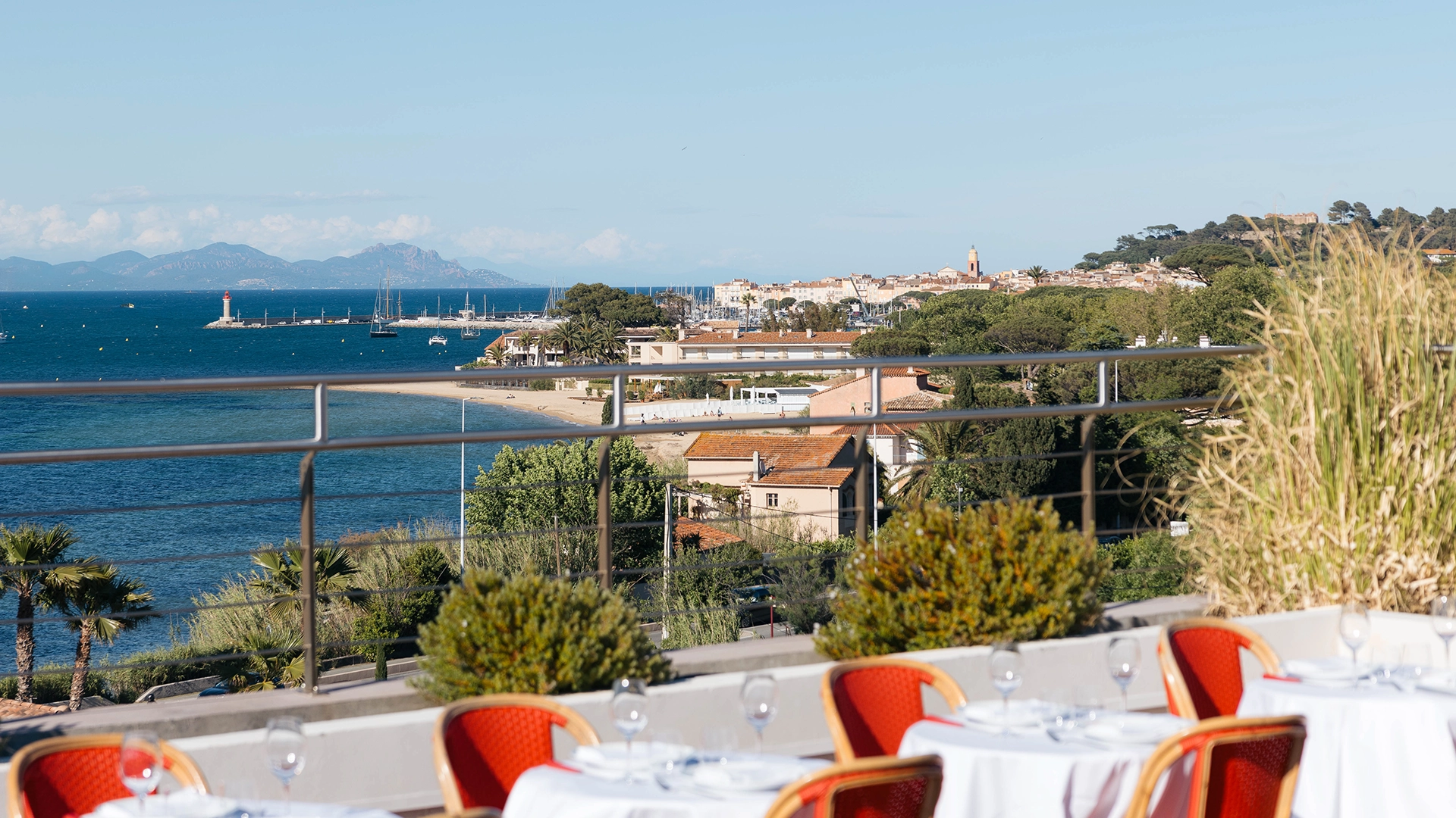 Terrasse avec tables dressées avec vue sur la plage, la mer, des bateaux et Saint-Tropez au loin.