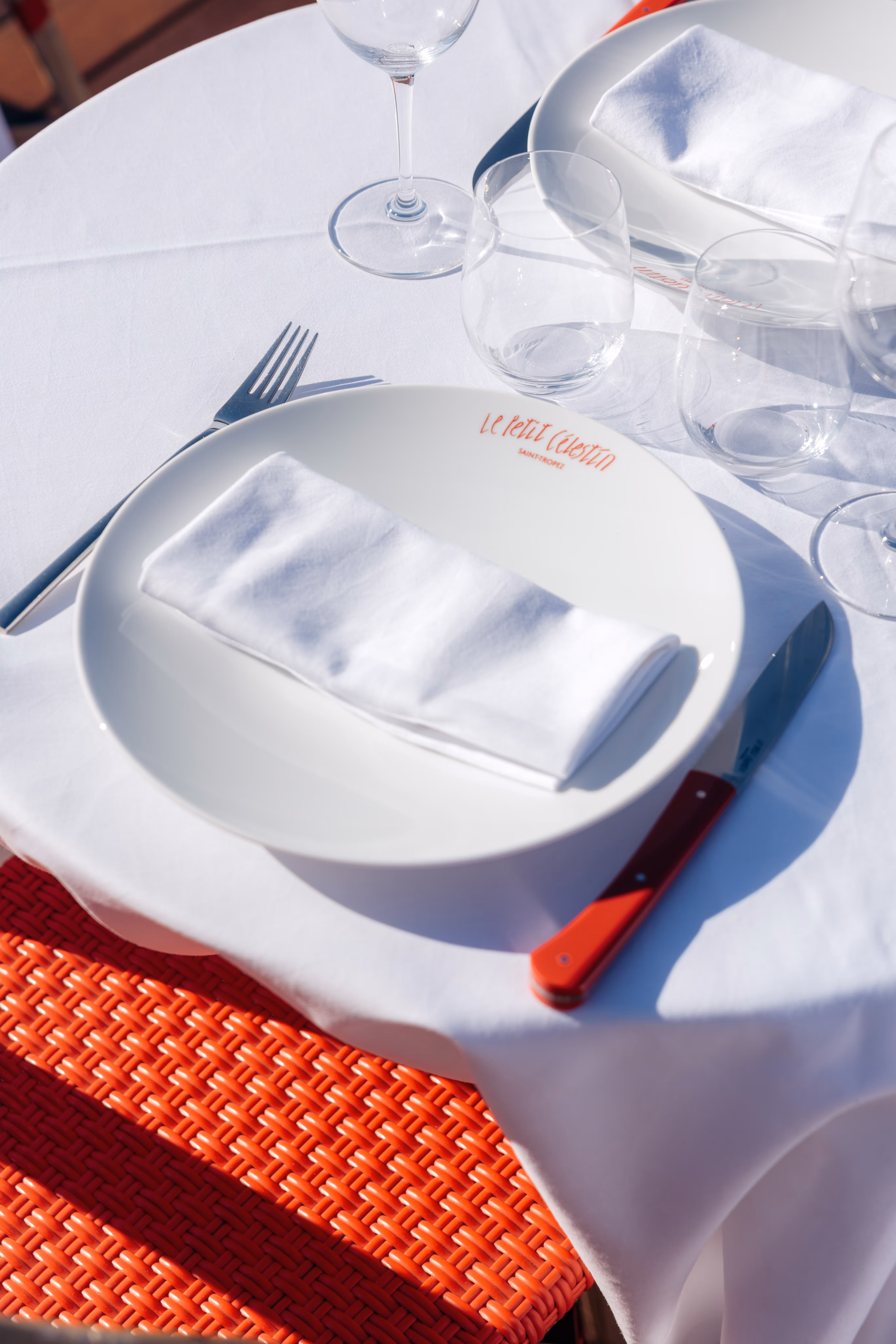 Table de restaurant avec assiette marquée Le Petit Celestin, serviette blanche, verre et couteau rouge.
