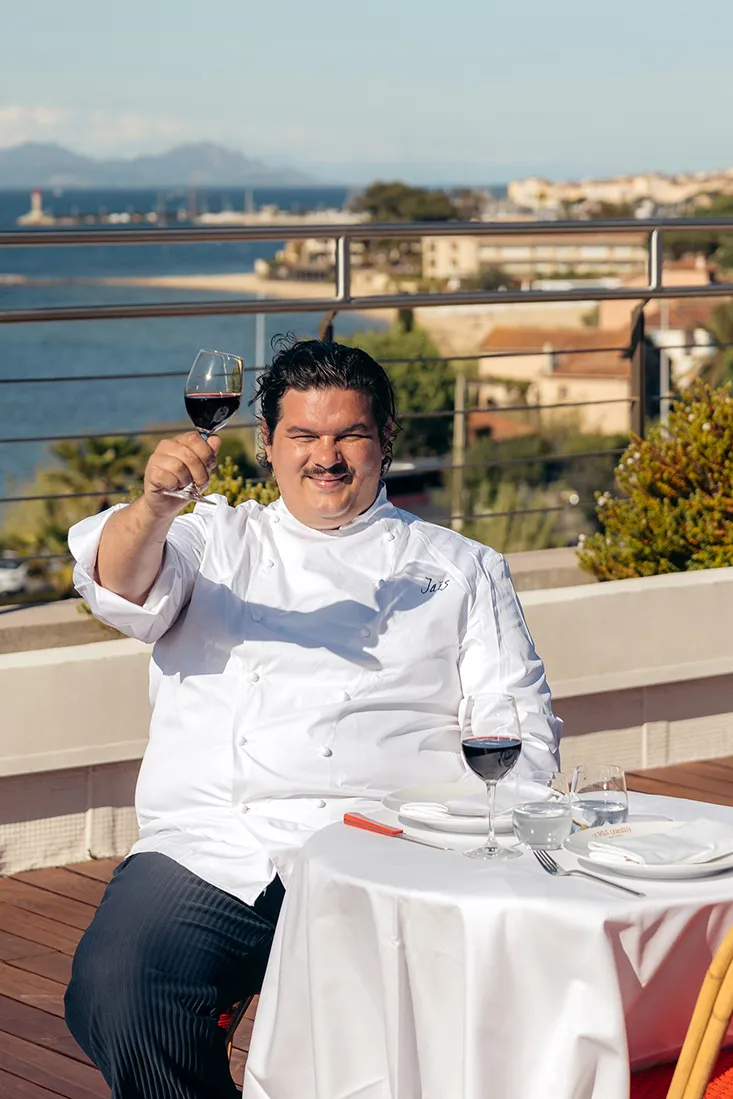Chef souriant en veste blanche levant un verre de vin rouge sur une terrasse avec vue sur l'eau.
