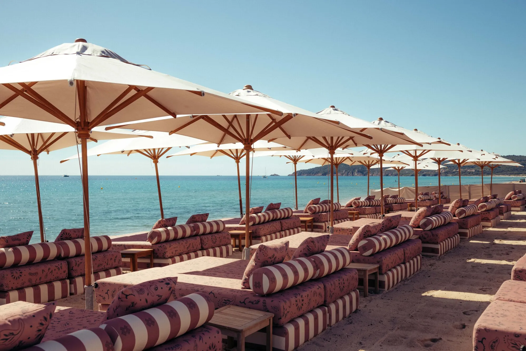 Lits de plage alignés avec coussins rayés et parasols blancs au bord de la mer sous un ciel bleu clair.