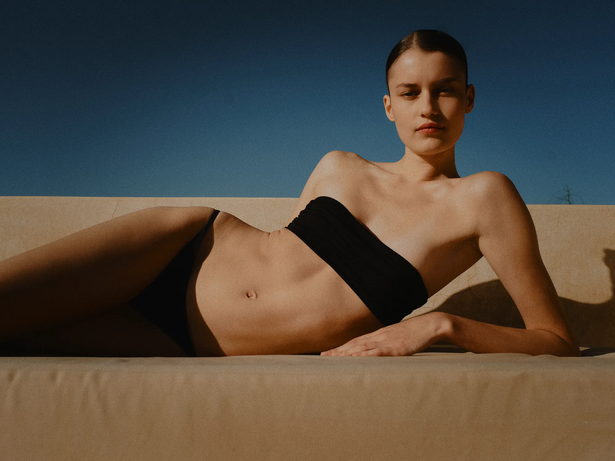 Femme allongée en bikini noir sur un canapé beige sous un ciel bleu dégagé.