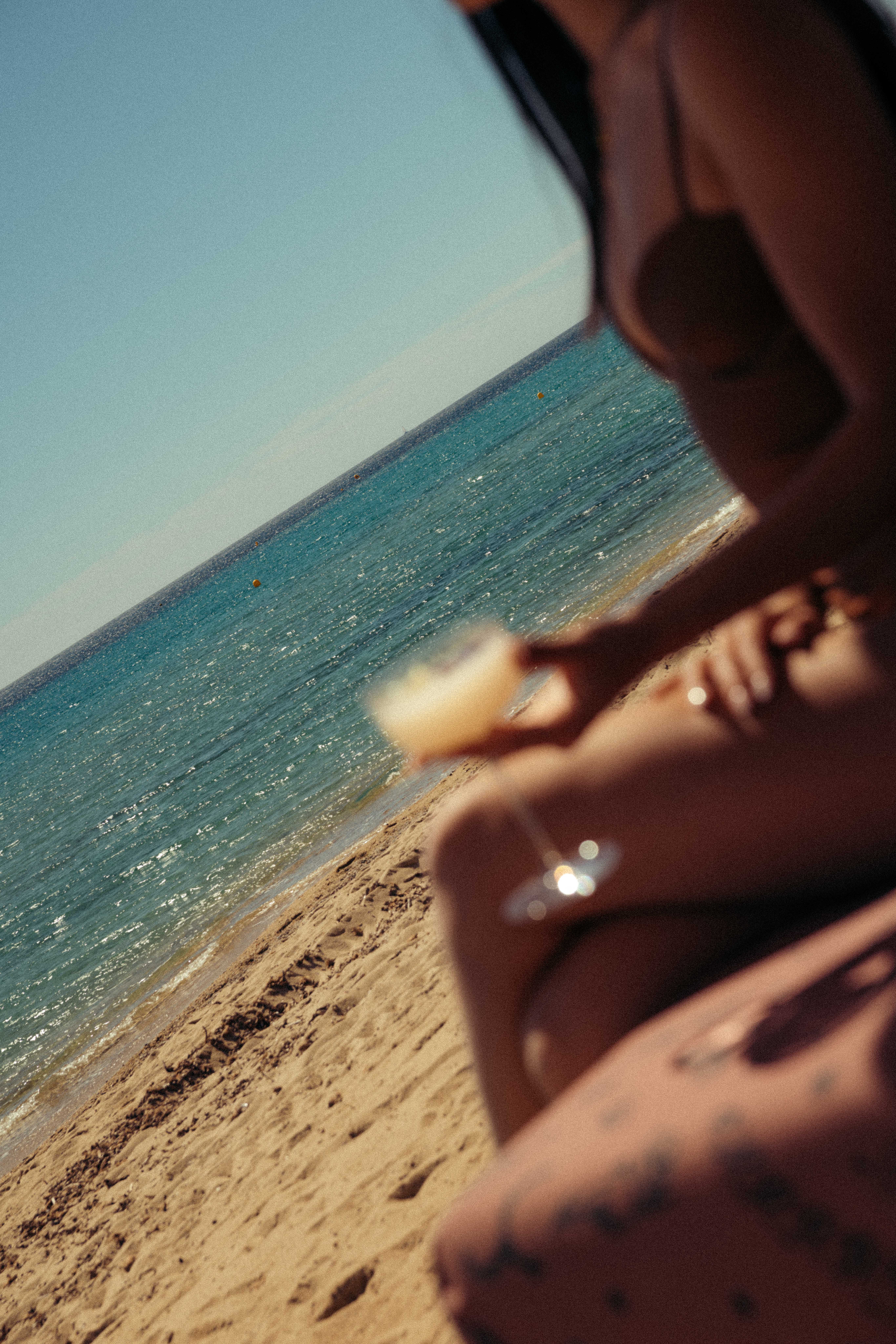 Femme en maillot de bain tenant un cocktail assise sur la plage près de la mer bleue.