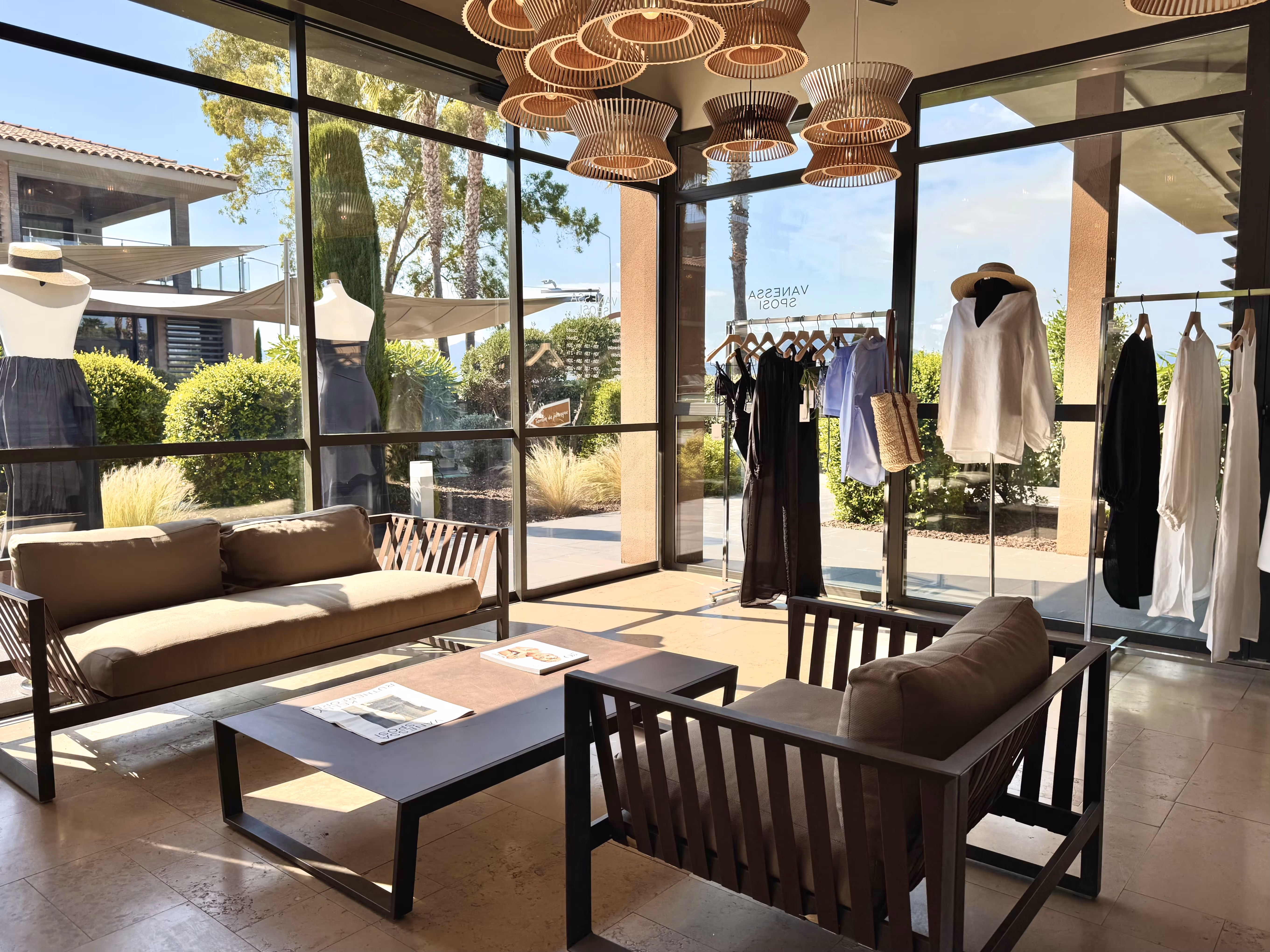 Coin salon avec canapés, table basse, mannequins vêtus et vêtements suspendus derrière une vitre.