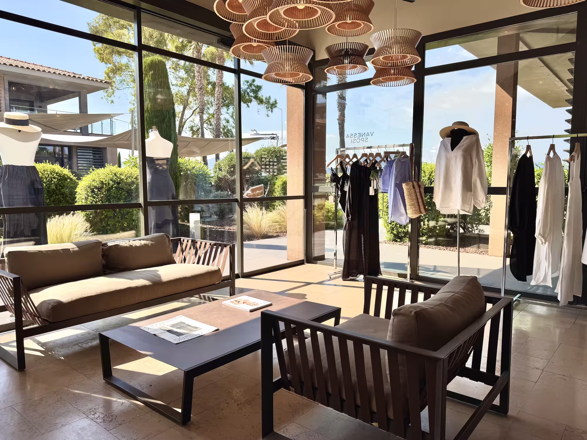 Coin salon avec canapés, table basse, mannequins vêtus et vêtements suspendus derrière une vitre.