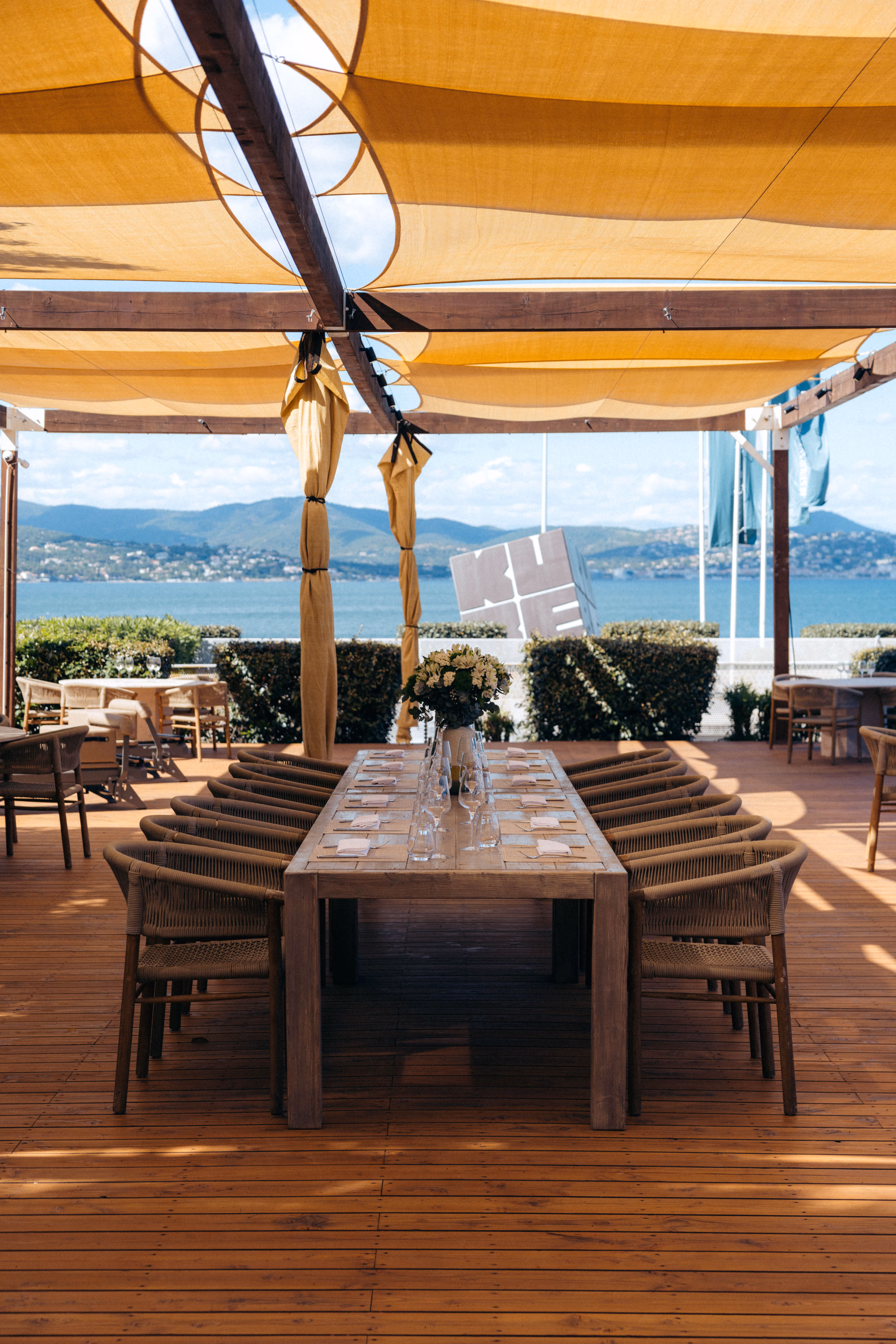 Table en bois dressée avec chaises sous un abri face à la mer et montagnes au loin.