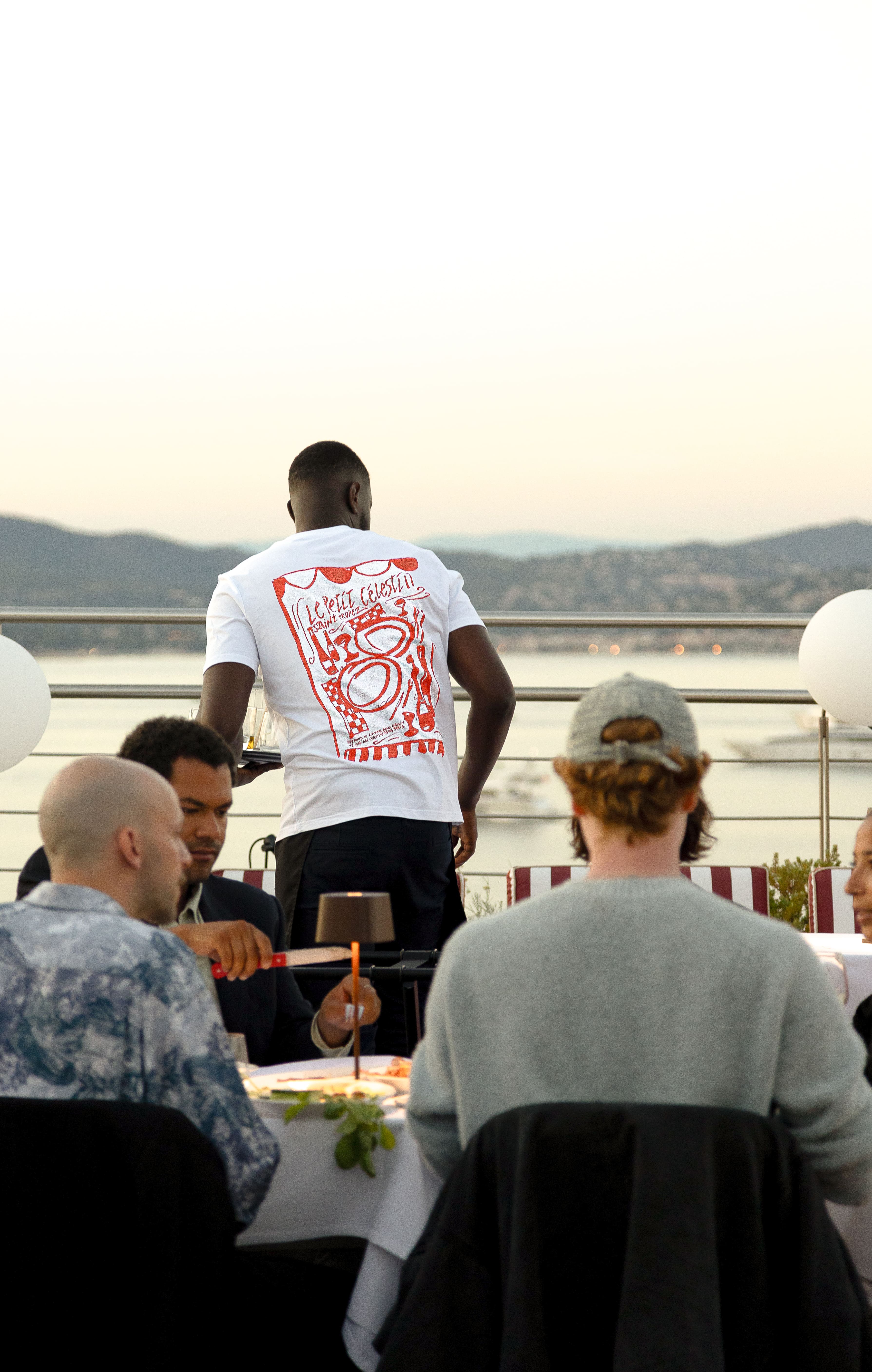 Serveur portant un t-shirt blanc avec un dessin rouge, servant des clients attablés sur une terrasse en soirée.