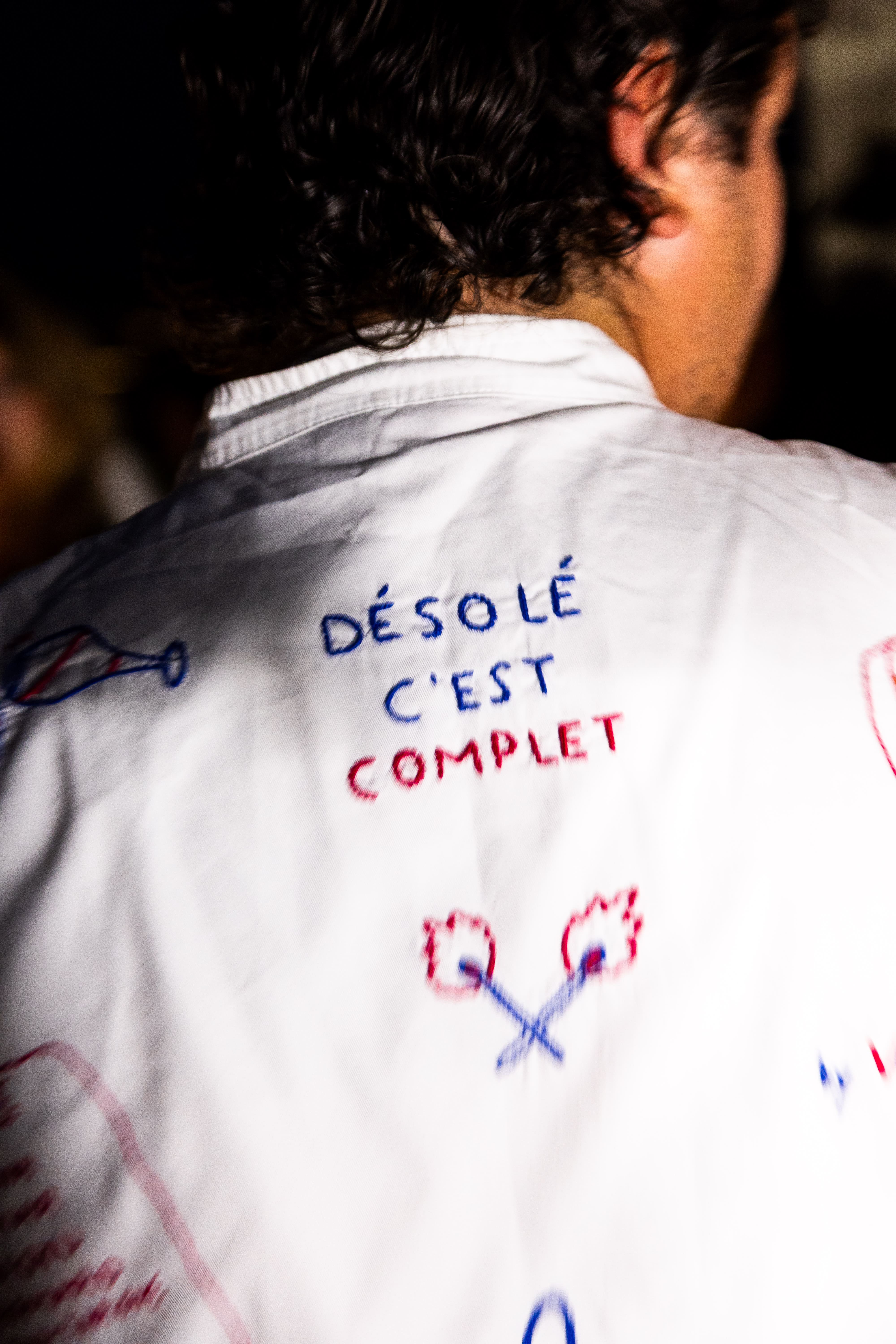 Dos d'une personne portant une chemise blanche avec texte brodé "Désolé c'est complet" et dessins.