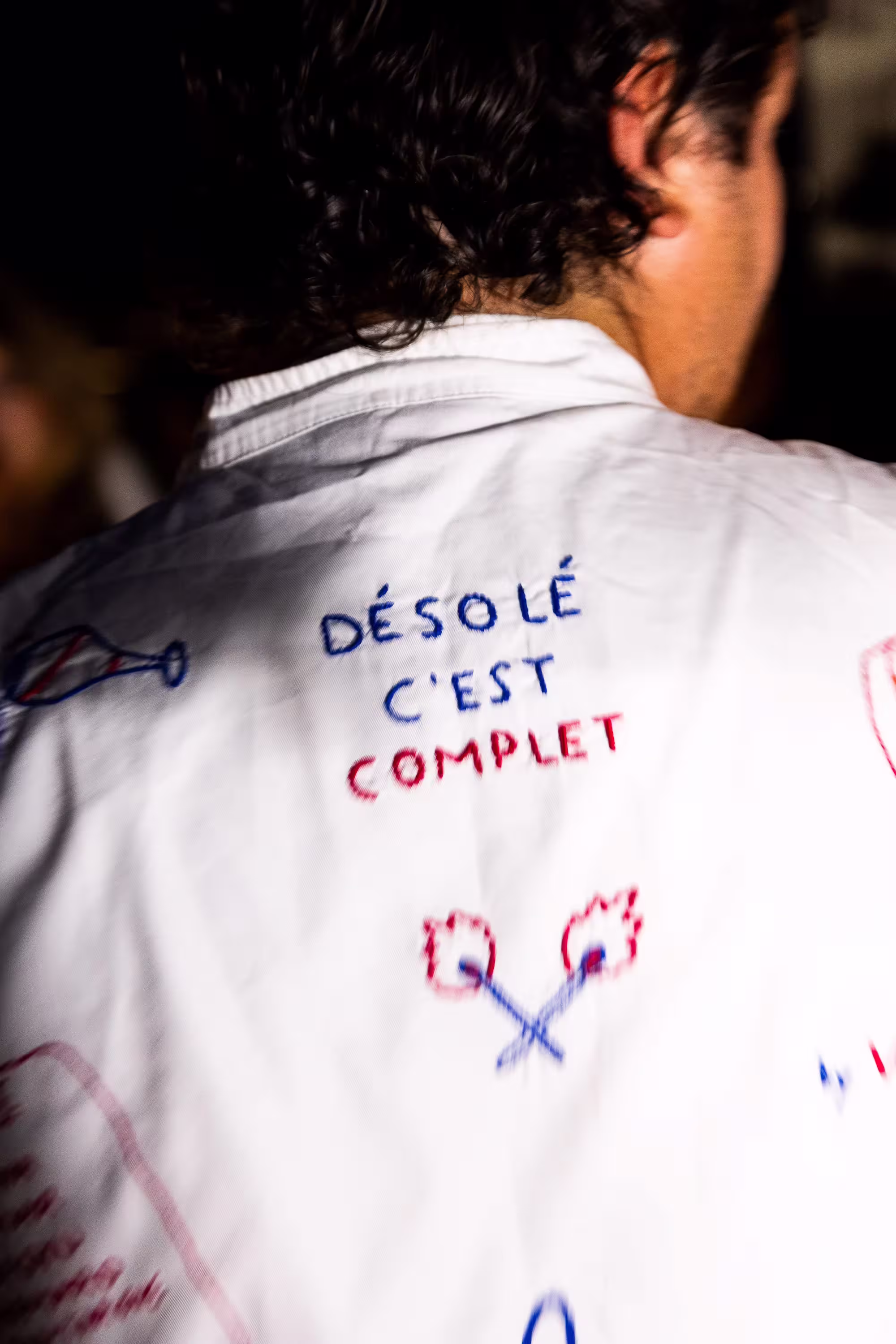 Dos d'une personne portant une chemise blanche avec texte brodé "Désolé c'est complet" et dessins.
