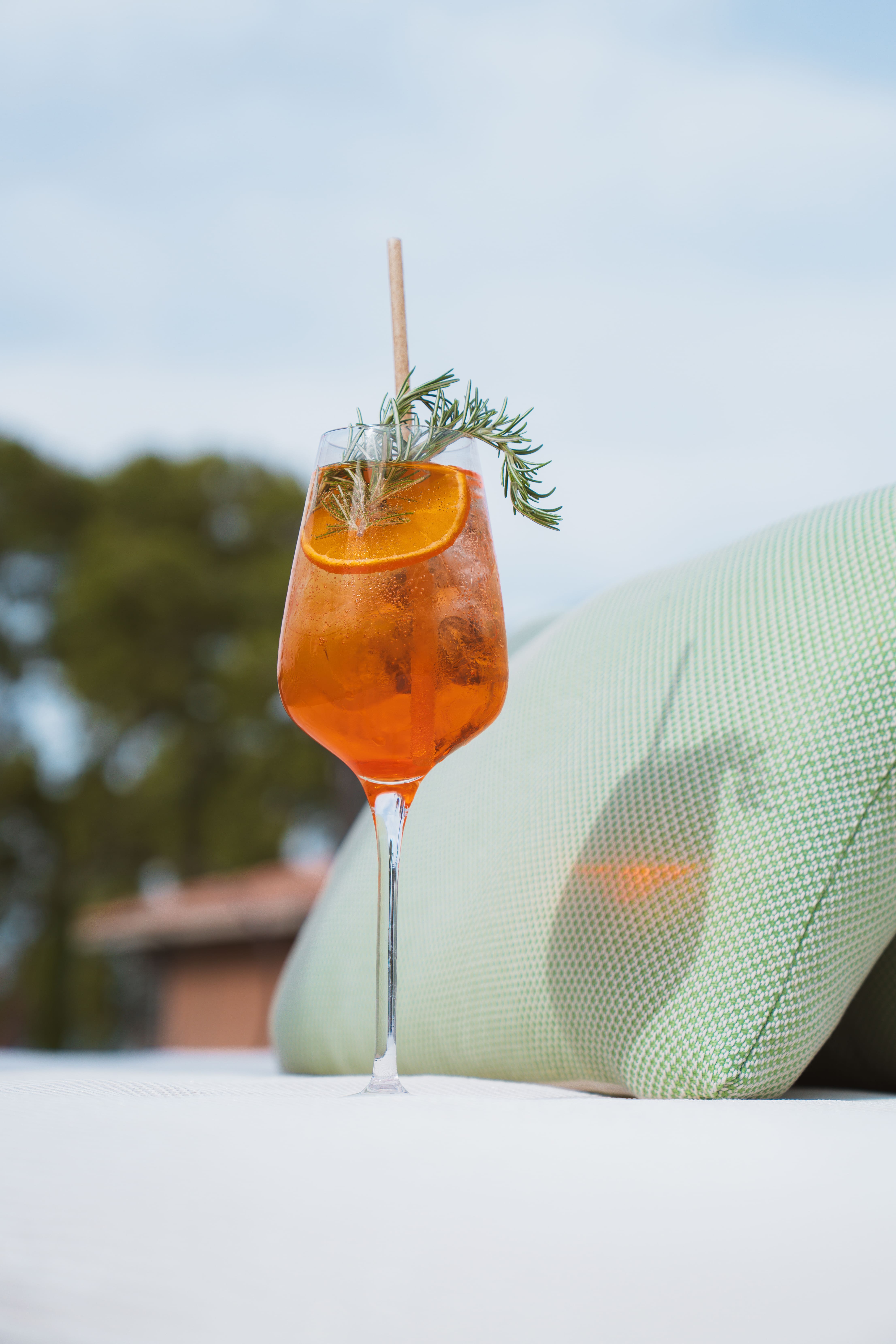 Verre à pied avec cocktail orange, tranche d'orange, branche de romarin et paille en extérieur.
