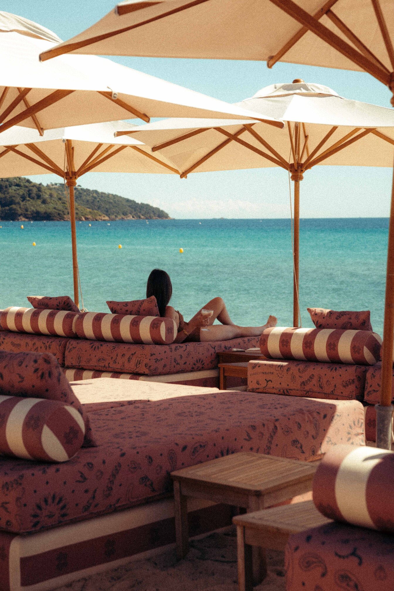 Femme se détendant sur un canapé rayé sous parasols, bord de mer turquoise et colline en arrière-plan.