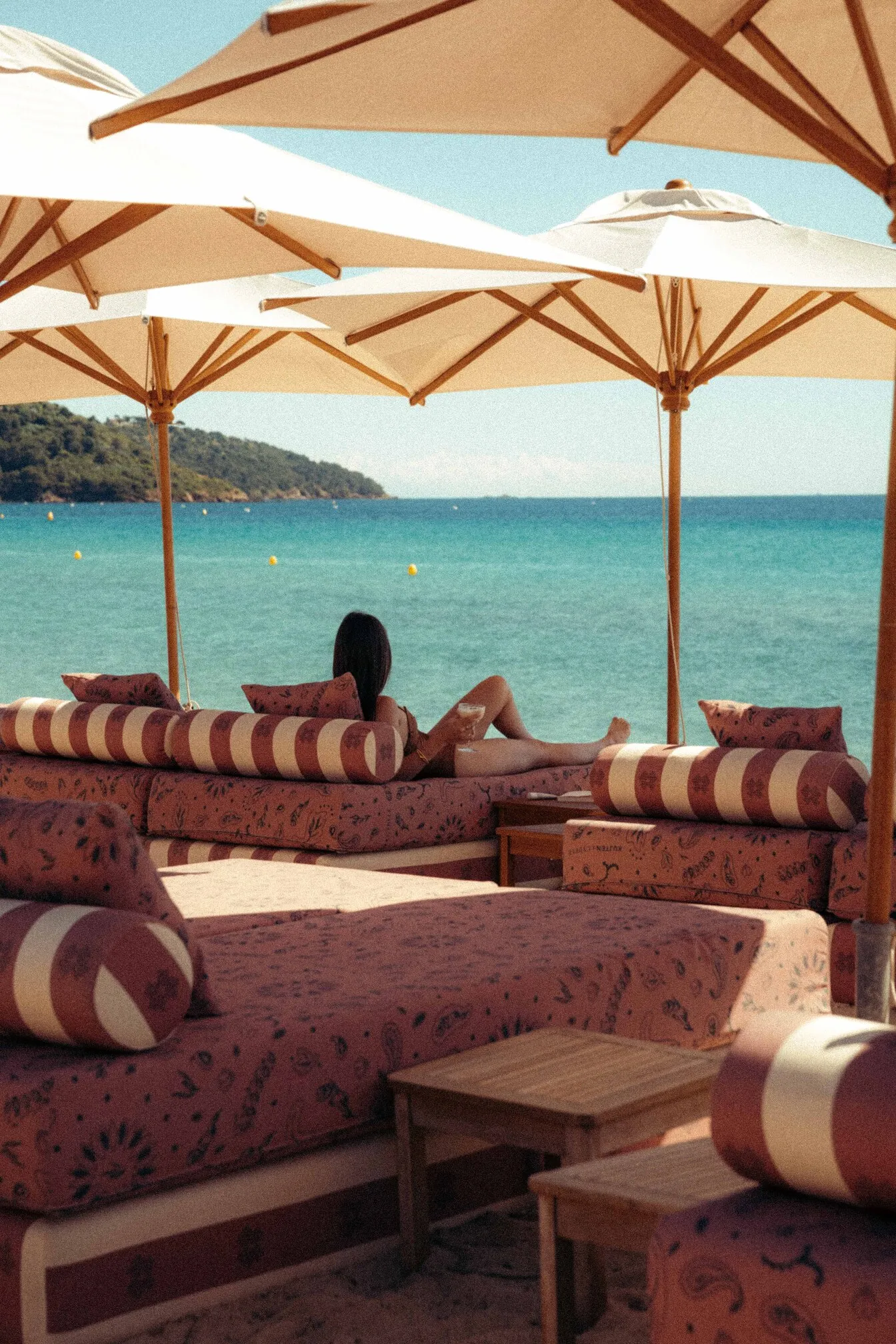 Femme se détendant sur un canapé rayé sous parasols, bord de mer turquoise et colline en arrière-plan.