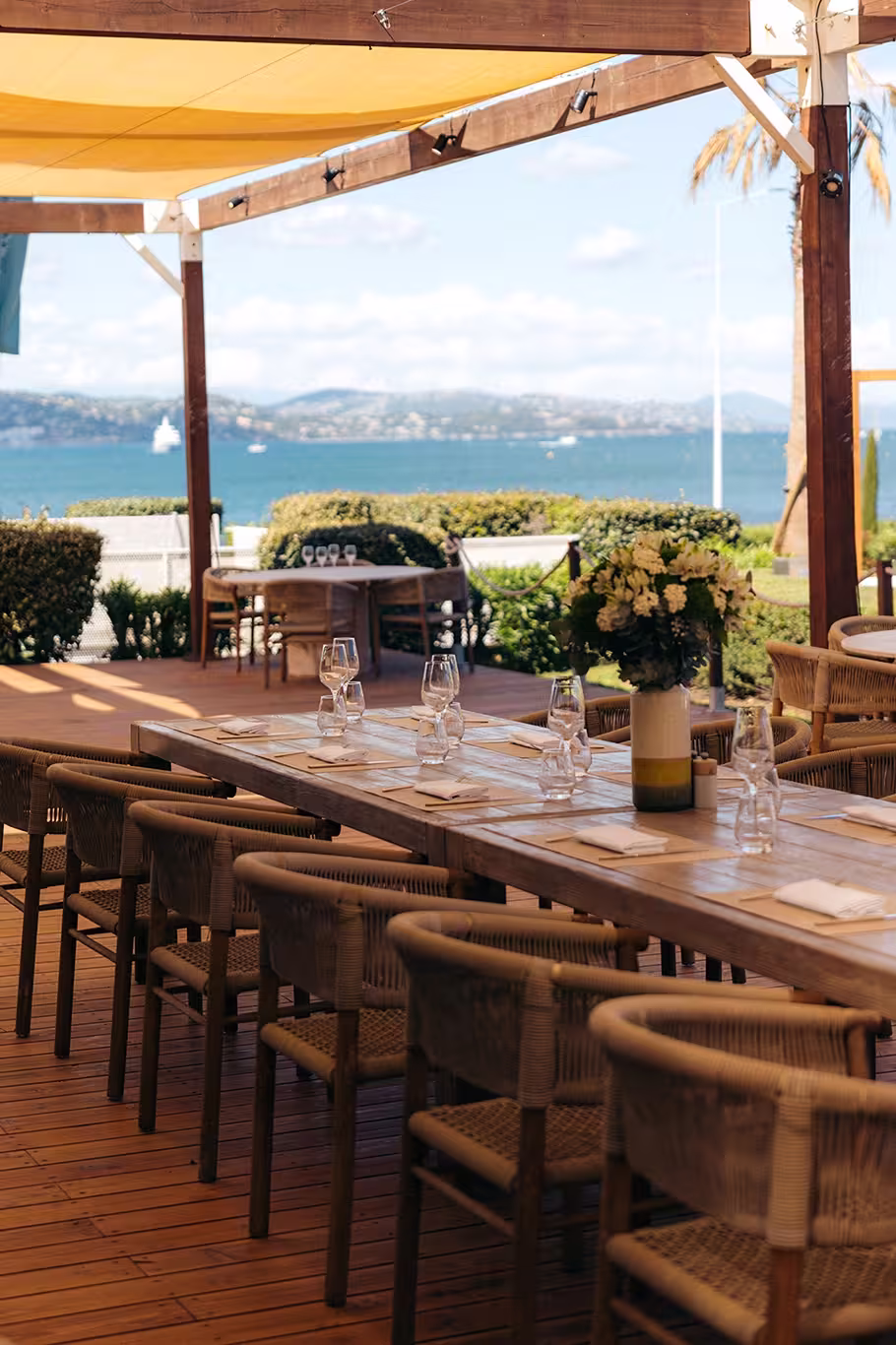 Terrasse de restaurant en bois avec table dressée, chaises en osier et vue sur mer au loin.