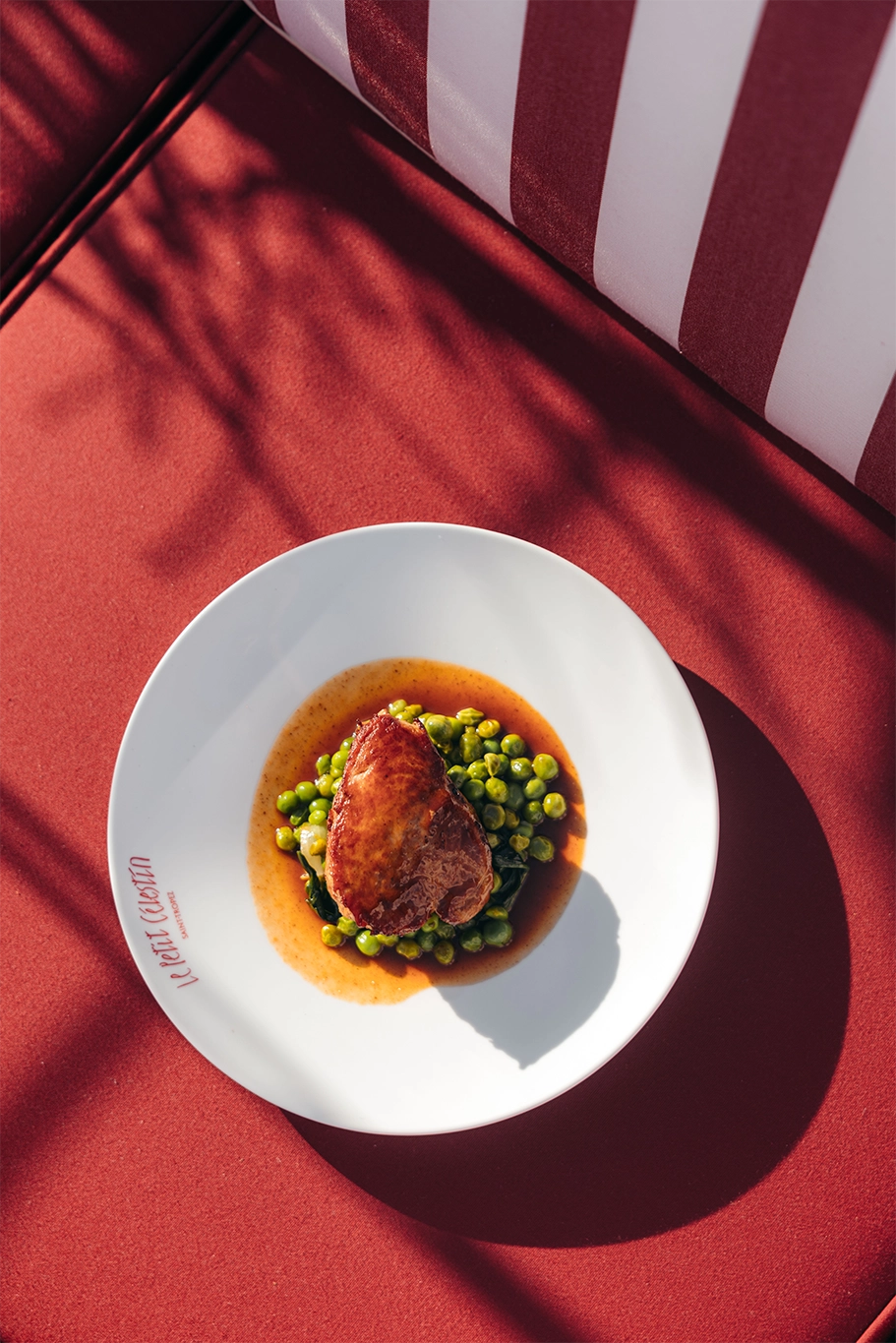 Assiette blanche avec un morceau de viande sur des petits pois verts et sauce sur un tissu rouge.