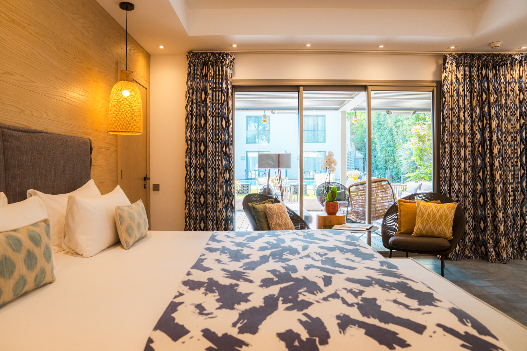 Chambre d'hôtel de luxe lumineuse avec jeté de lit aux motifs abstraits bleus, rideaux élégants et grande baie vitrée ouvrant sur une terrasse ensoleillée.