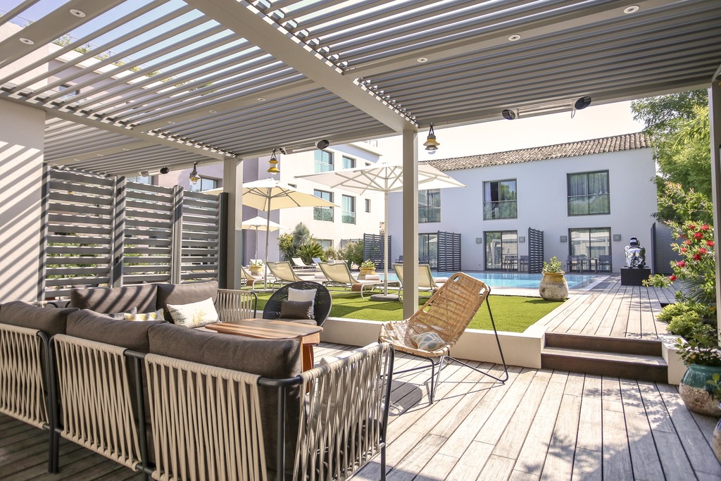 Espace détente extérieur avec salon de jardin moderne sous une pergola bioclimatique, vue sur la piscine de l'hôtel et façade de villa méditerranéenne