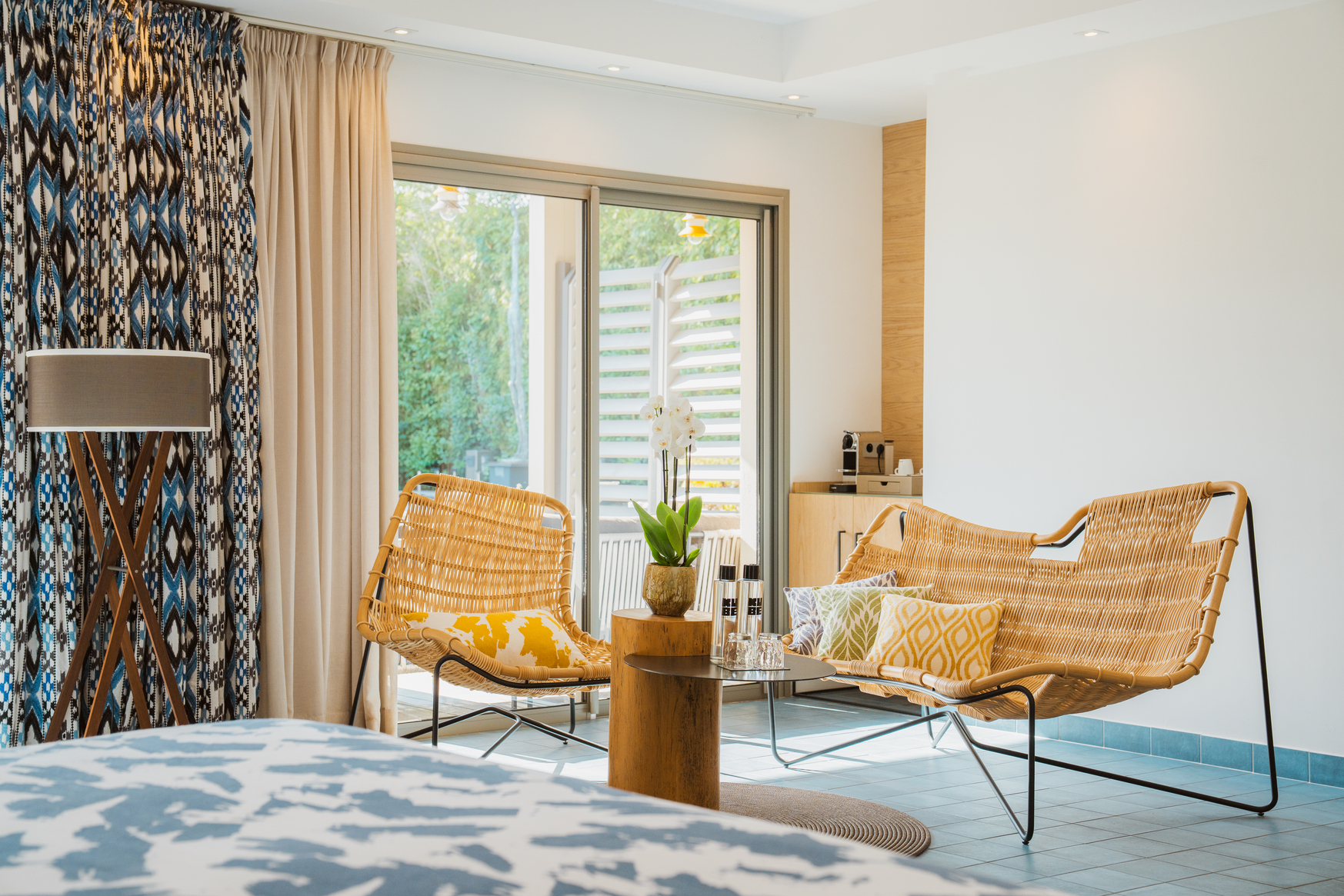 Espace salon dans une chambre style Ibiza avec fauteuils en rotin, rideaux à motifs ethniques et vue sur une terrasse arborée.