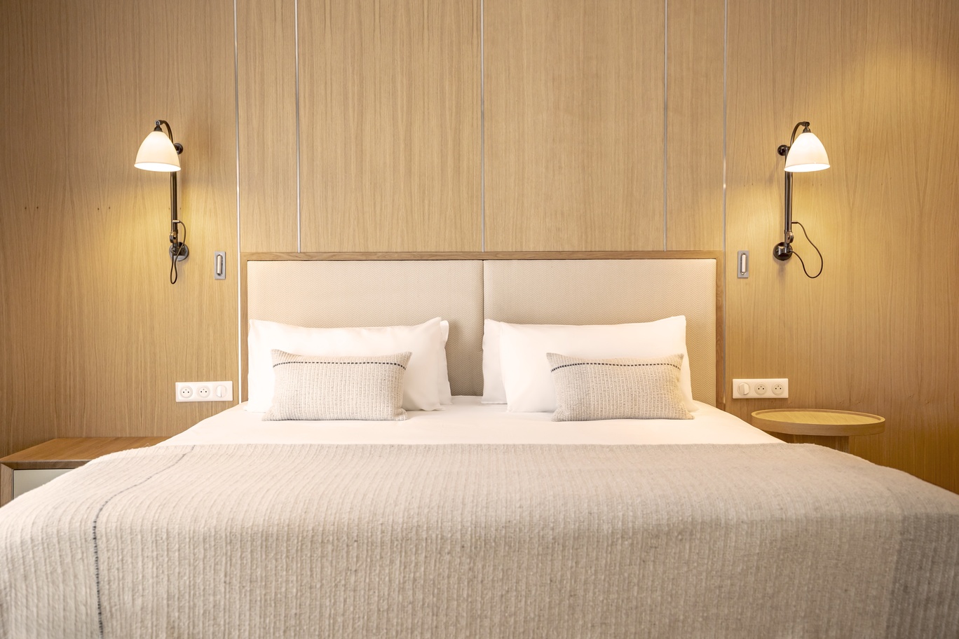 Chambre au design naturel avec tête de lit monumentale en bois clair, appliques murales vintage et linge de lit blanc épuré.