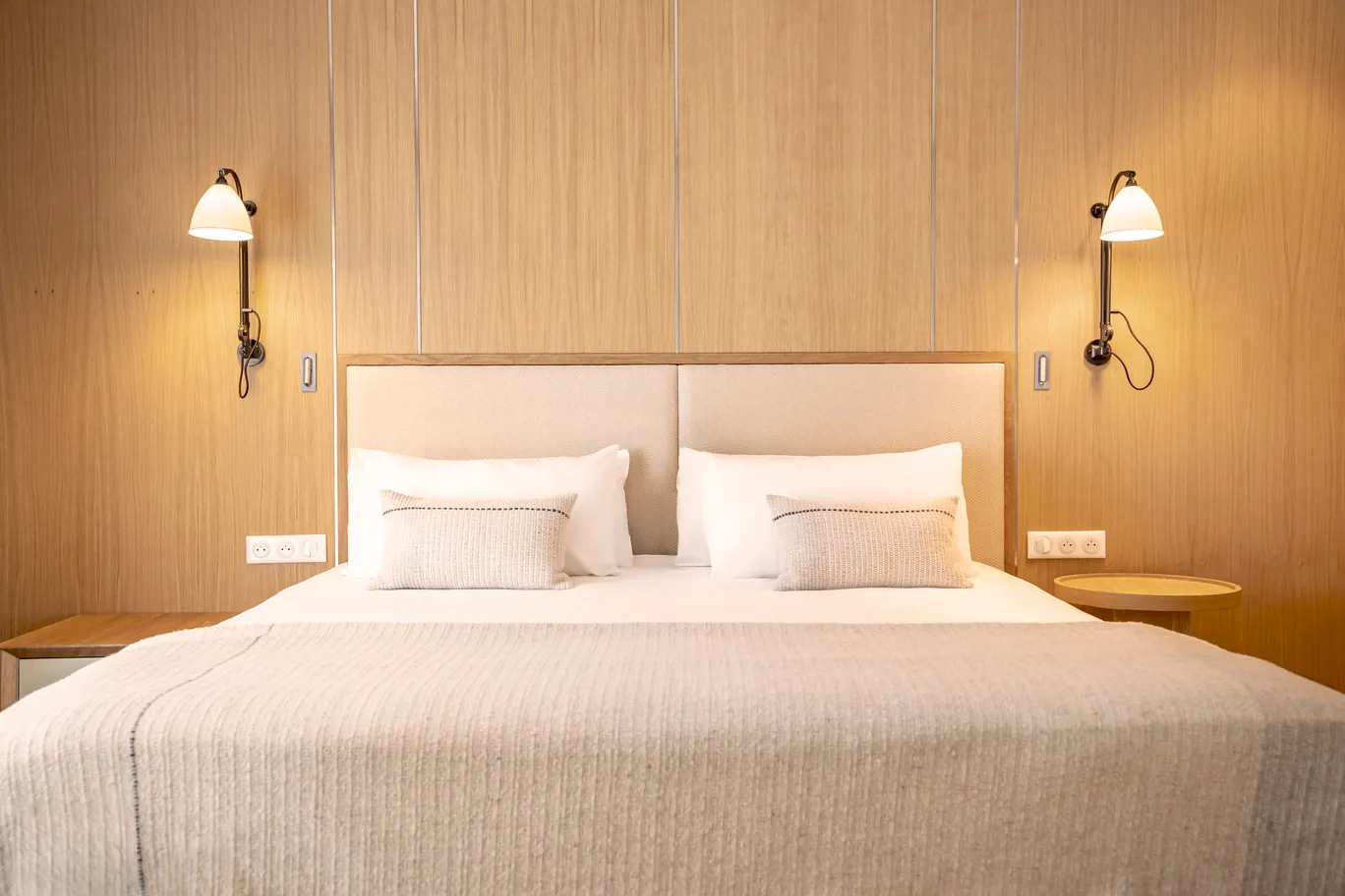 Chambre au design naturel avec tête de lit monumentale en bois clair, appliques murales vintage et linge de lit blanc épuré.