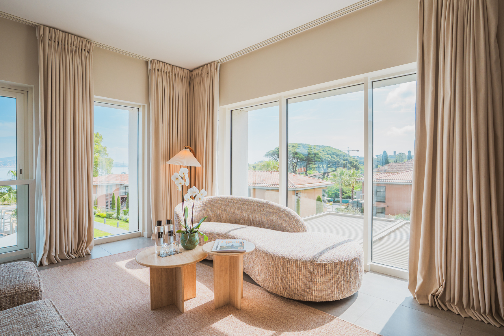 Espace salon d'une suite de luxe avec canapé design organique, tables en bois clair et vue panoramique sur le jardin et la mer.