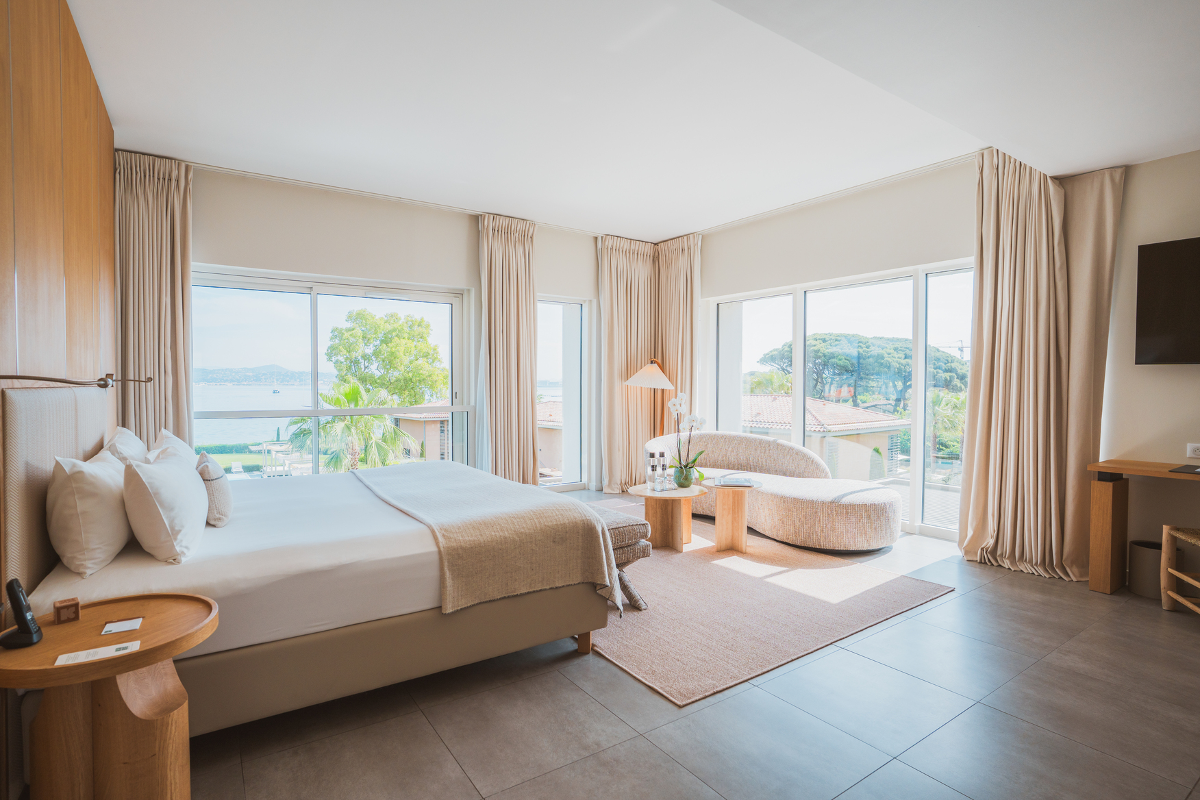 Suite Junior lumineuse avec design naturel, grand lit double face aux baies vitrées et salon incurvé contemporain avec vue mer