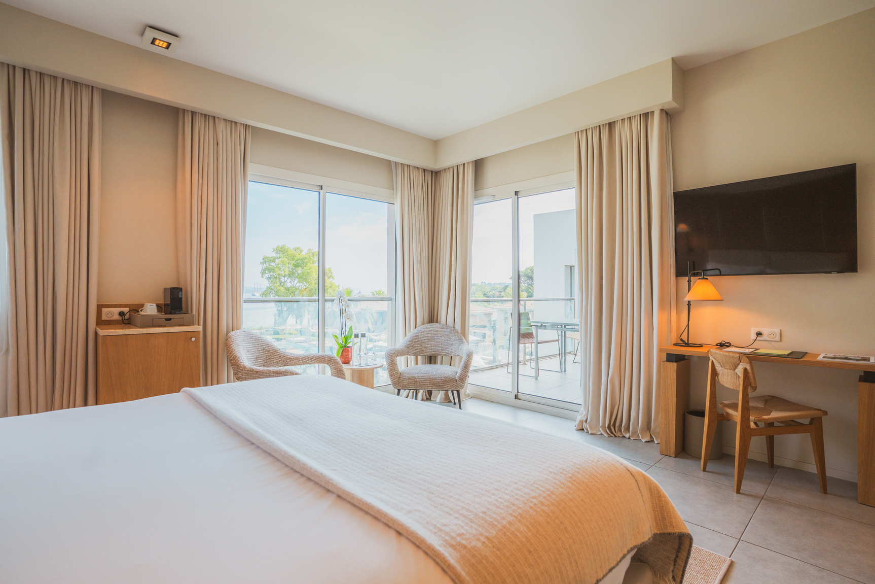 Salon privé de suite Prestige avec tables basses organiques, parquet clair et balcon filant offrant une vue sur la piscine et la nature.