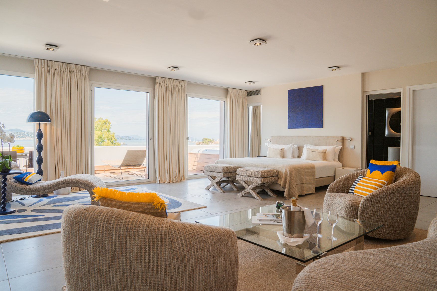 Salon de suite avec mobilier design contemporain, chaise longue ergonomique et grandes ouvertures sur un balcon filant face à l'océan.