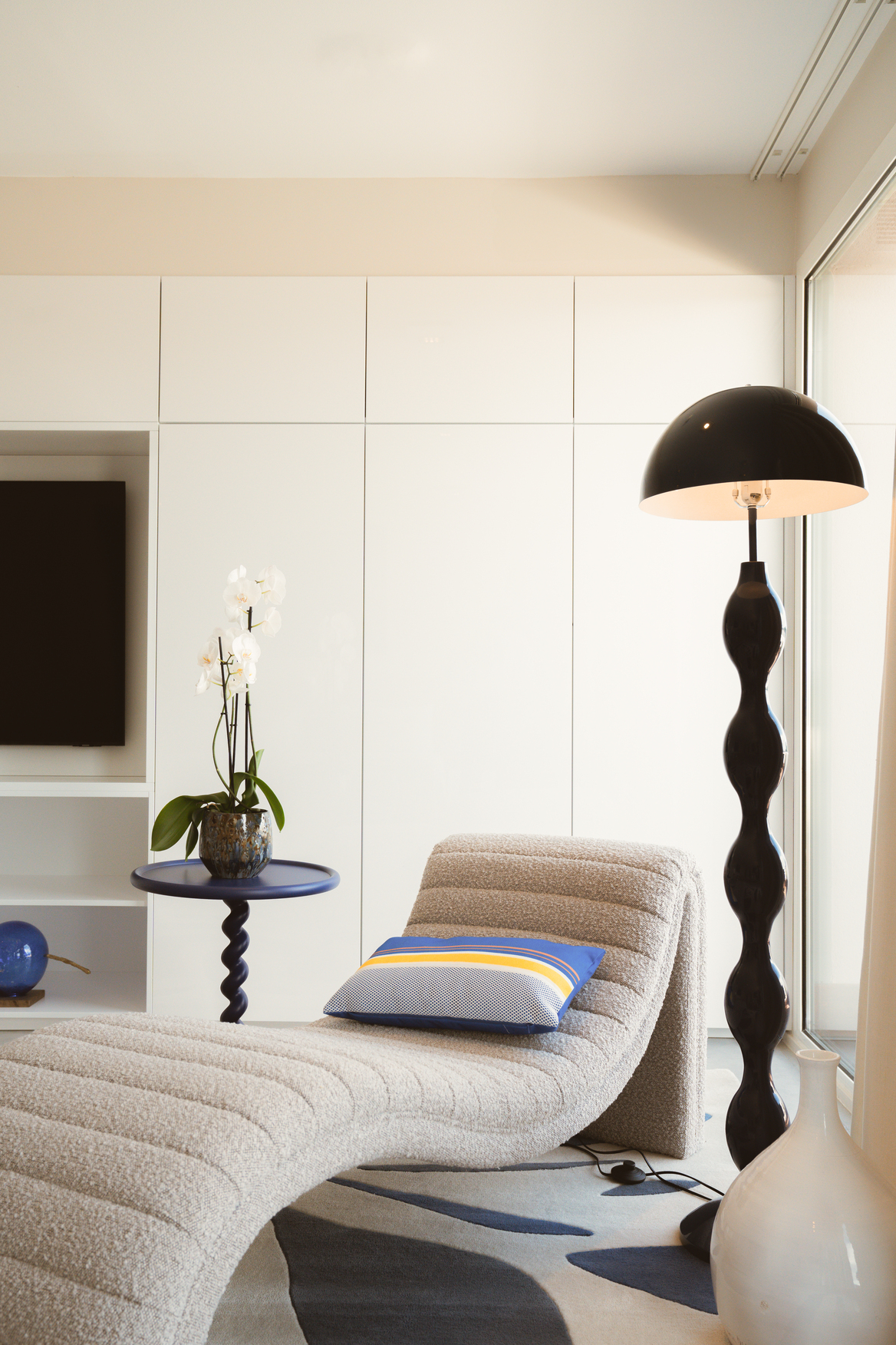 Espace détente intérieur avec chaise longue design, tapis aux motifs marins et salon lumineux prolongé par une terrasse en bois.