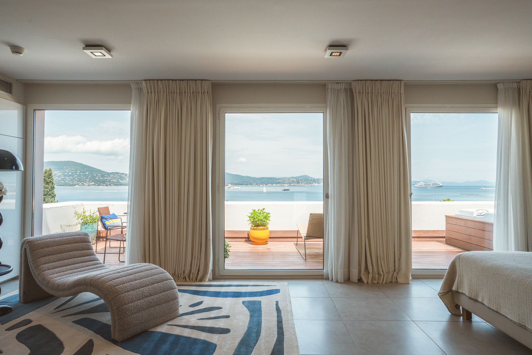 Salon de suite avec mobilier design contemporain, chaise longue ergonomique et grandes ouvertures sur un balcon filant face à l'océan.