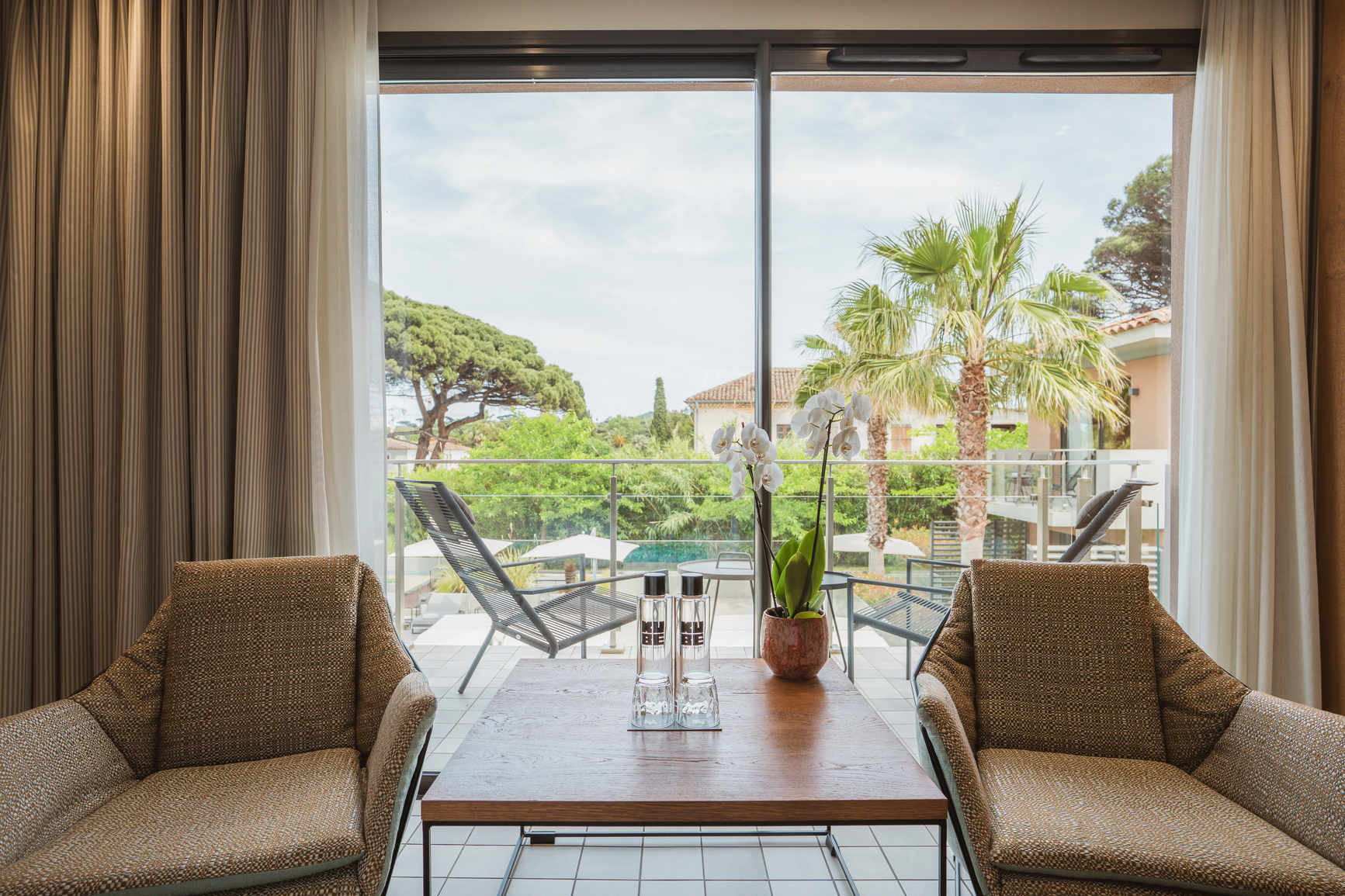 Espace salon dans une chambre style Ibiza avec fauteuils en rotin, rideaux à motifs ethniques et vue sur une terrasse arborée.