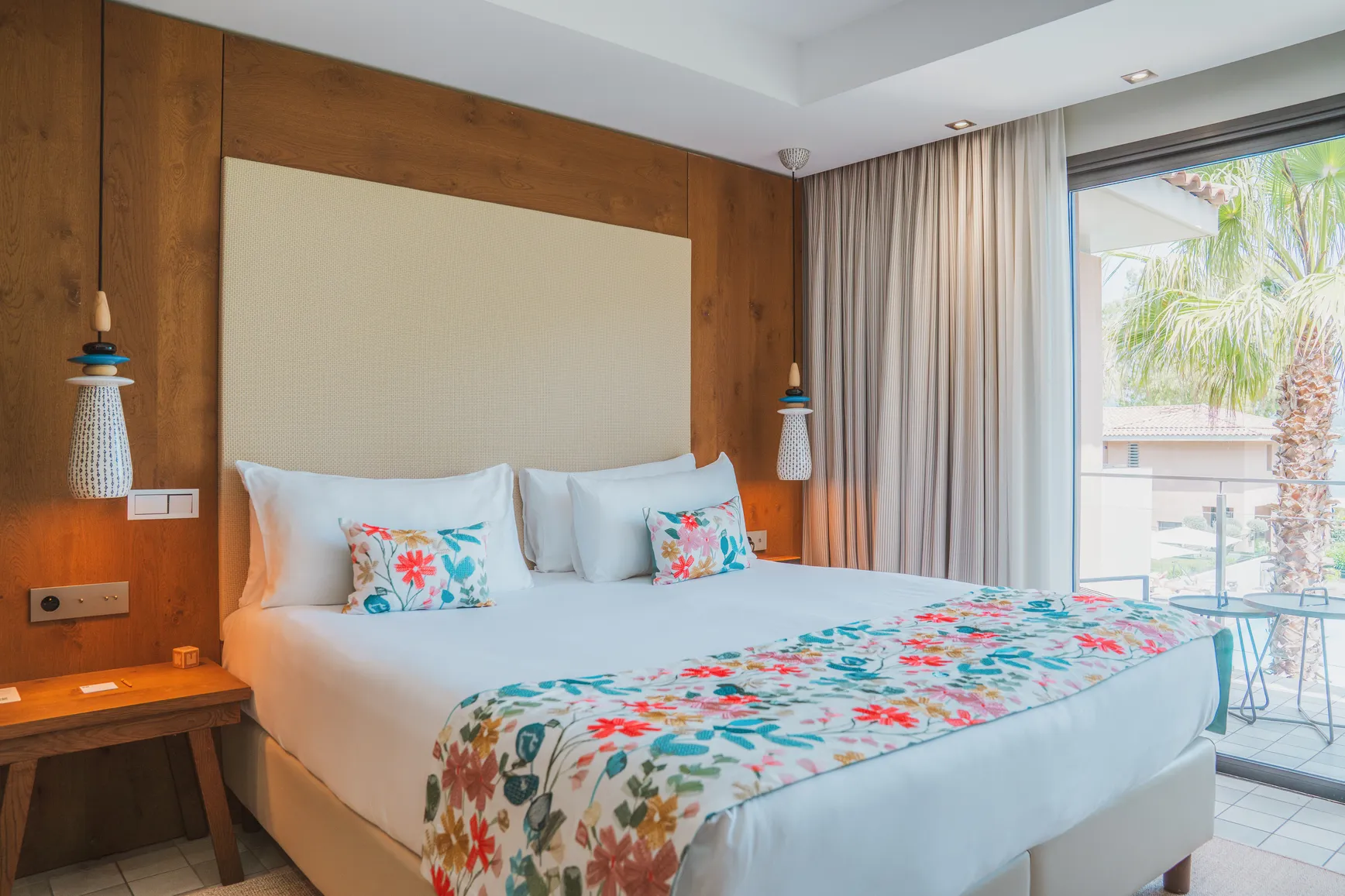 Chambre Deluxe Wood avec tête de lit en bois massif, literie aux motifs floraux et vue sur le jardin méditerranéen.