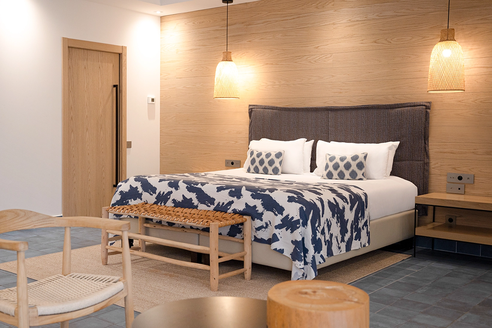 Chambre style Ibiza chic avec tête de lit en bois clair, literie aux motifs graphiques bleus et lampe suspendue en fibres naturelles.