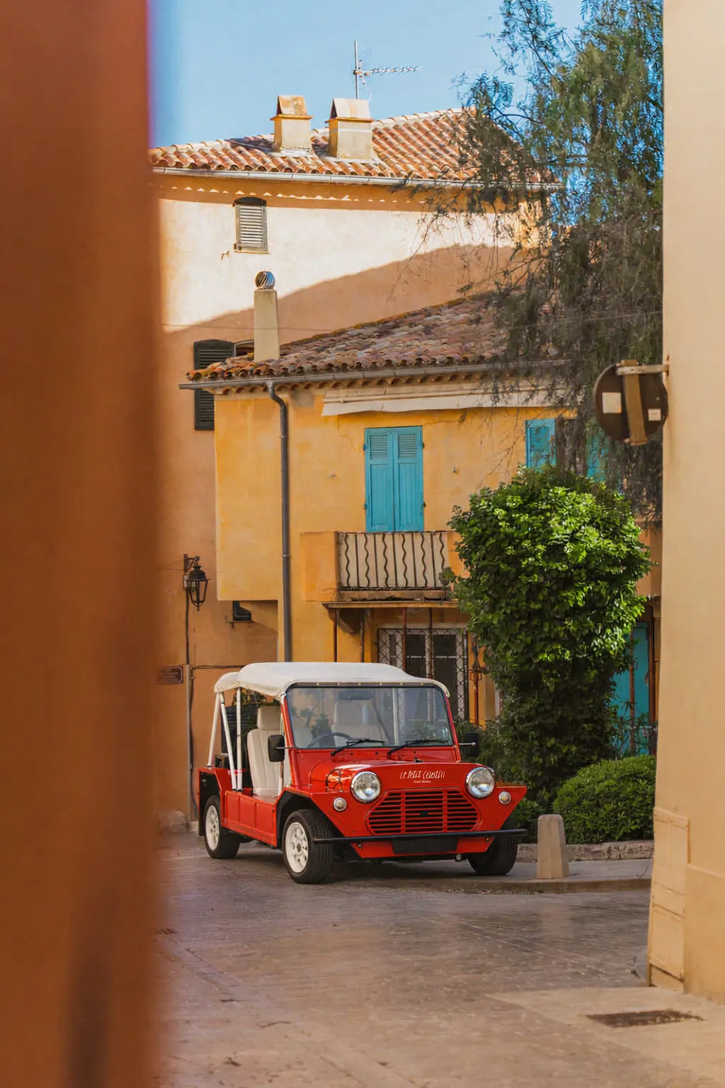 Voiturette de style Mini Moke rouge garée dans une ruelle ensoleillée aux façades provençales