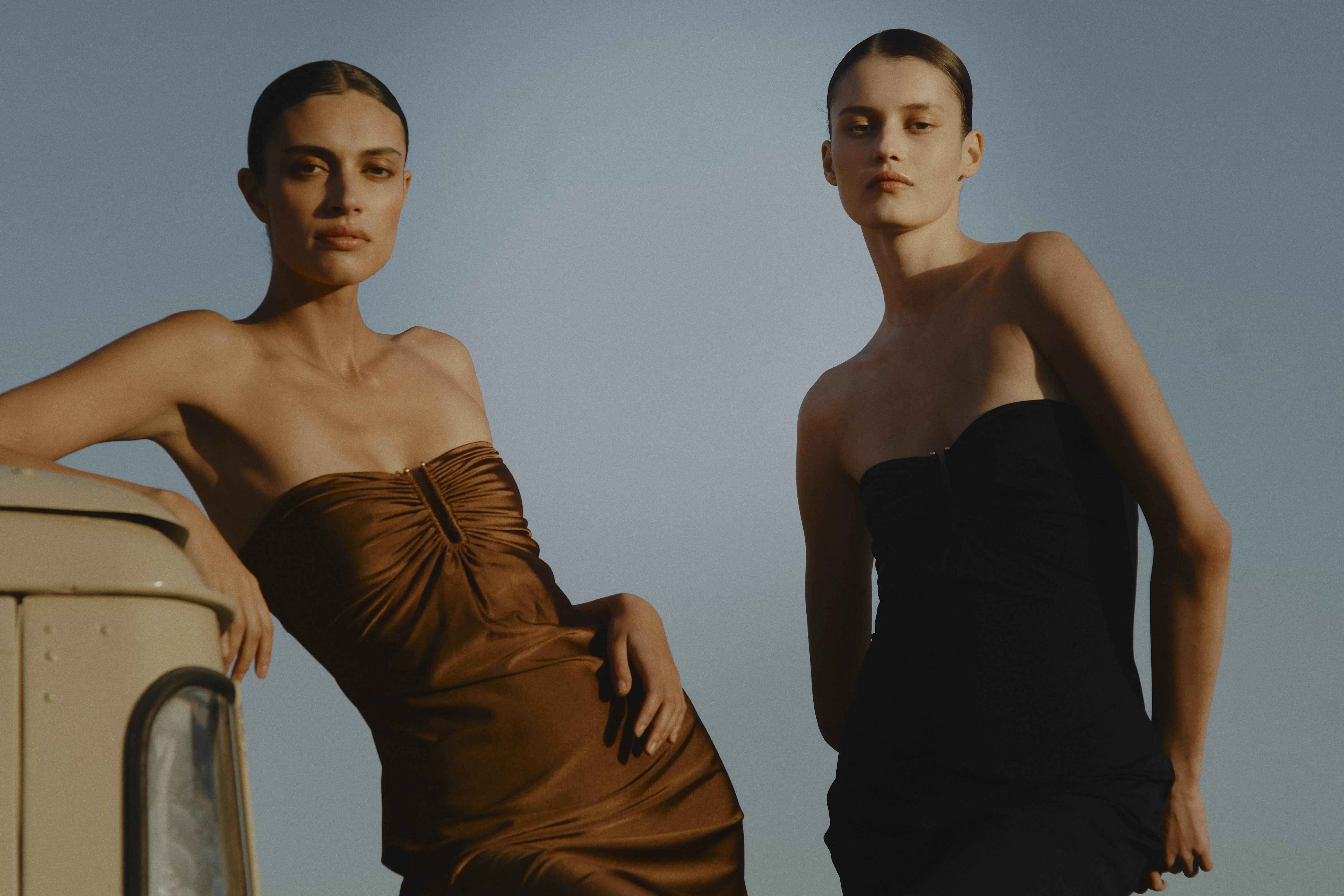Deux mannequins posent en extérieur avec des robes bustier drapées marron et noire signées Vanessa Sposi.