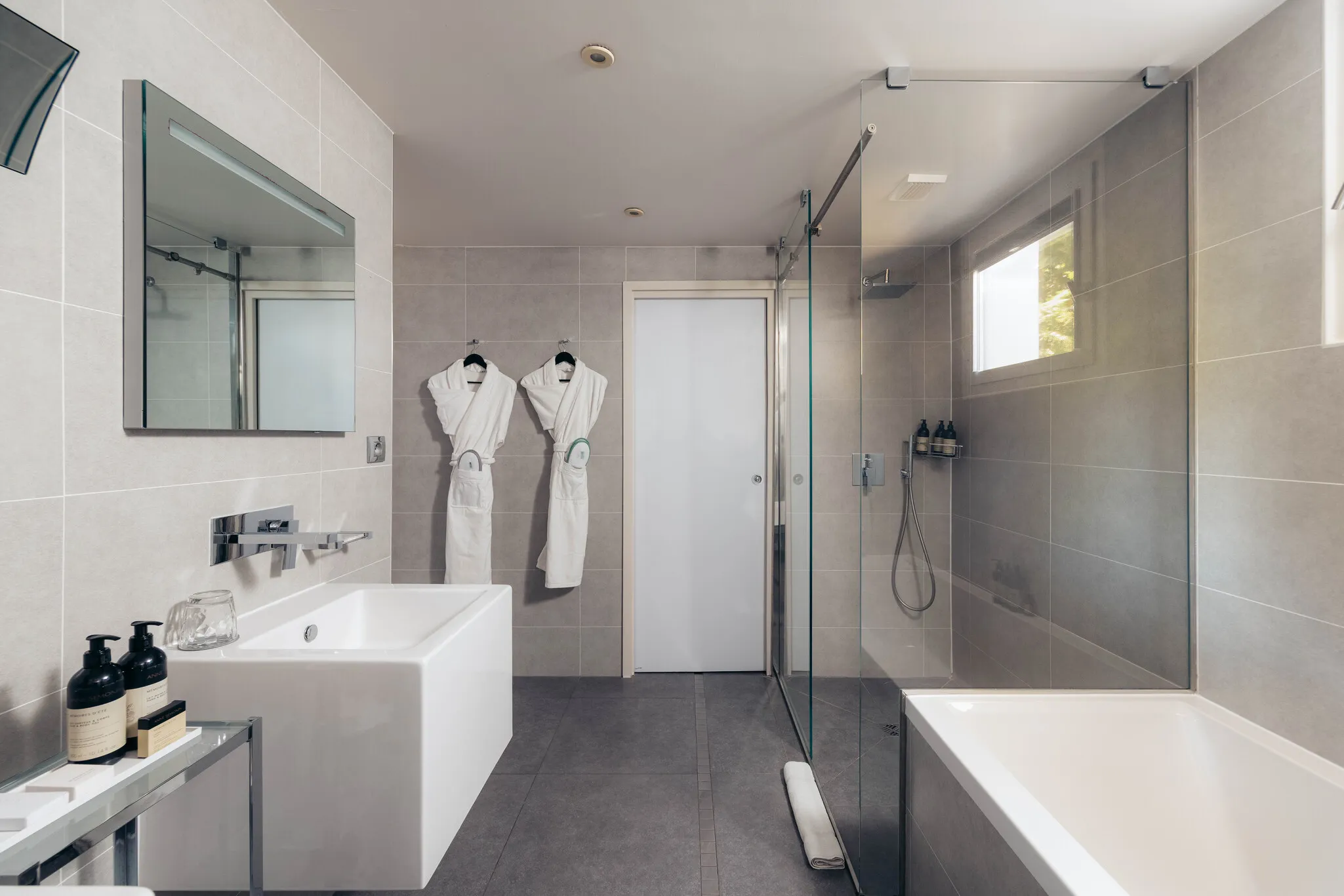 Grande salle de bain moderne avec douche à l'italienne vitrée, meuble vasque et peignoirs dans la suite Junior Natural.