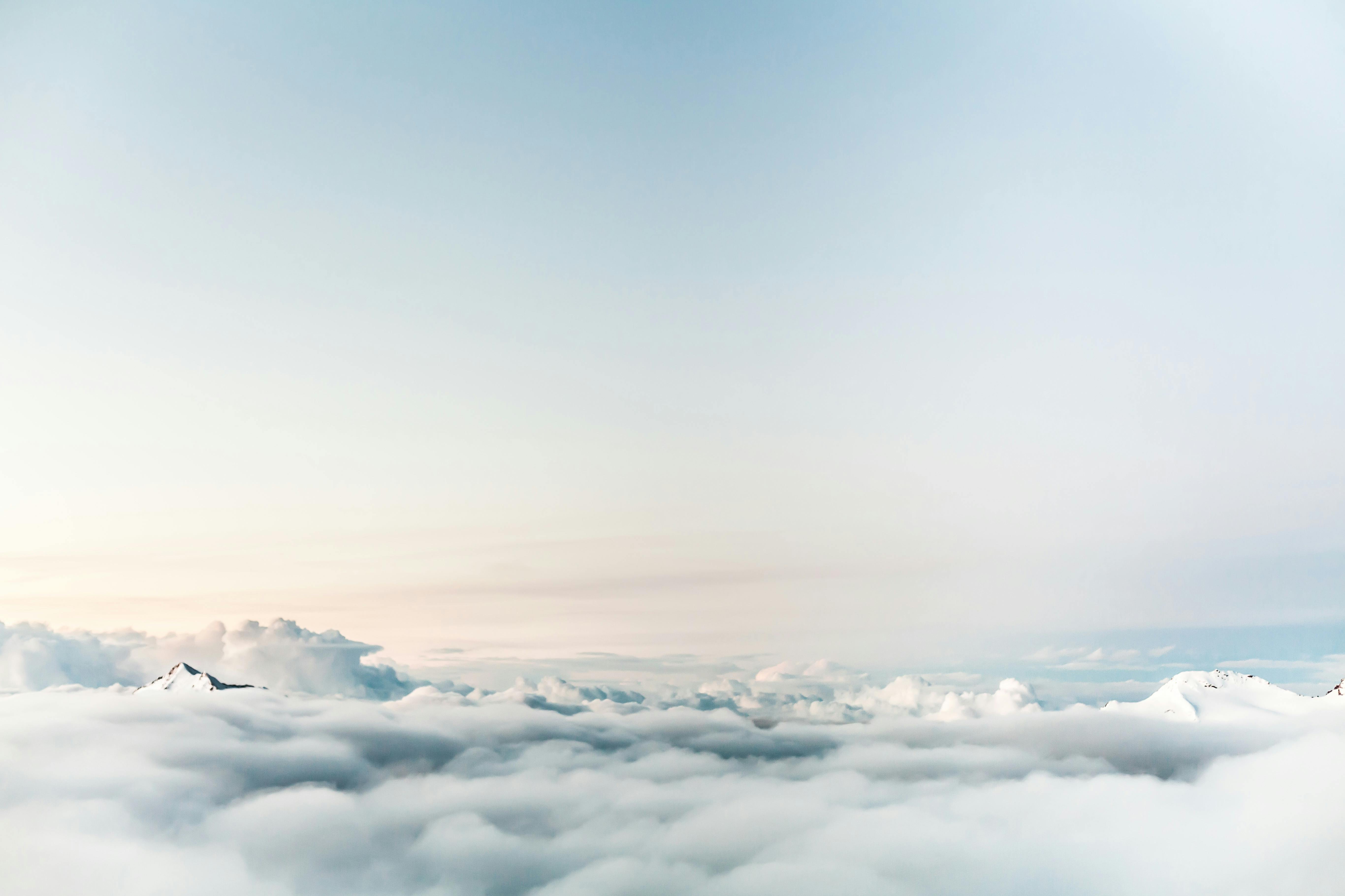 Snowy mountain peaks rising through a thick layer of clouds under a pale blue sky.