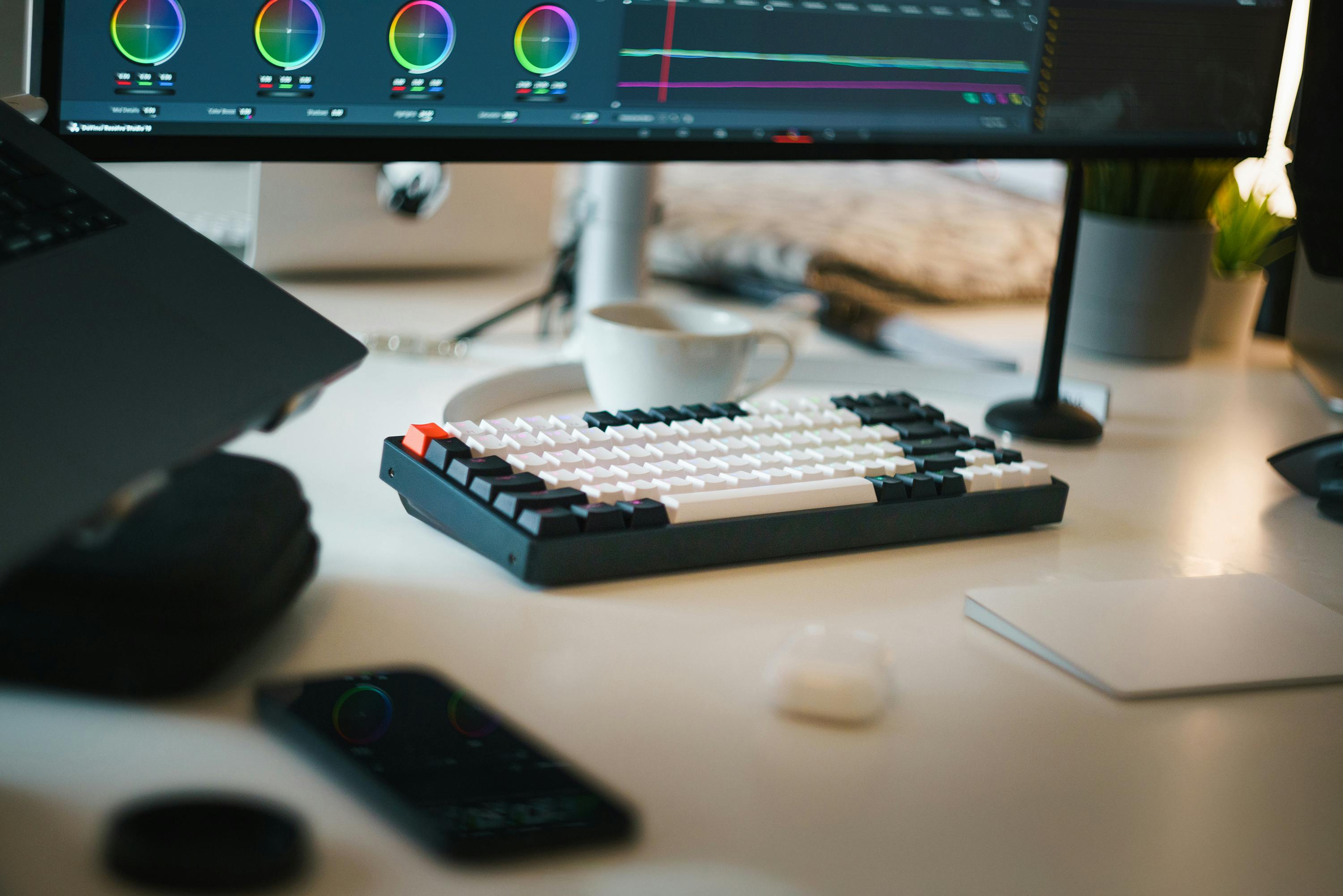 Mechanical keyboard with white and black keys on a desk with a cup, monitor, and plants.