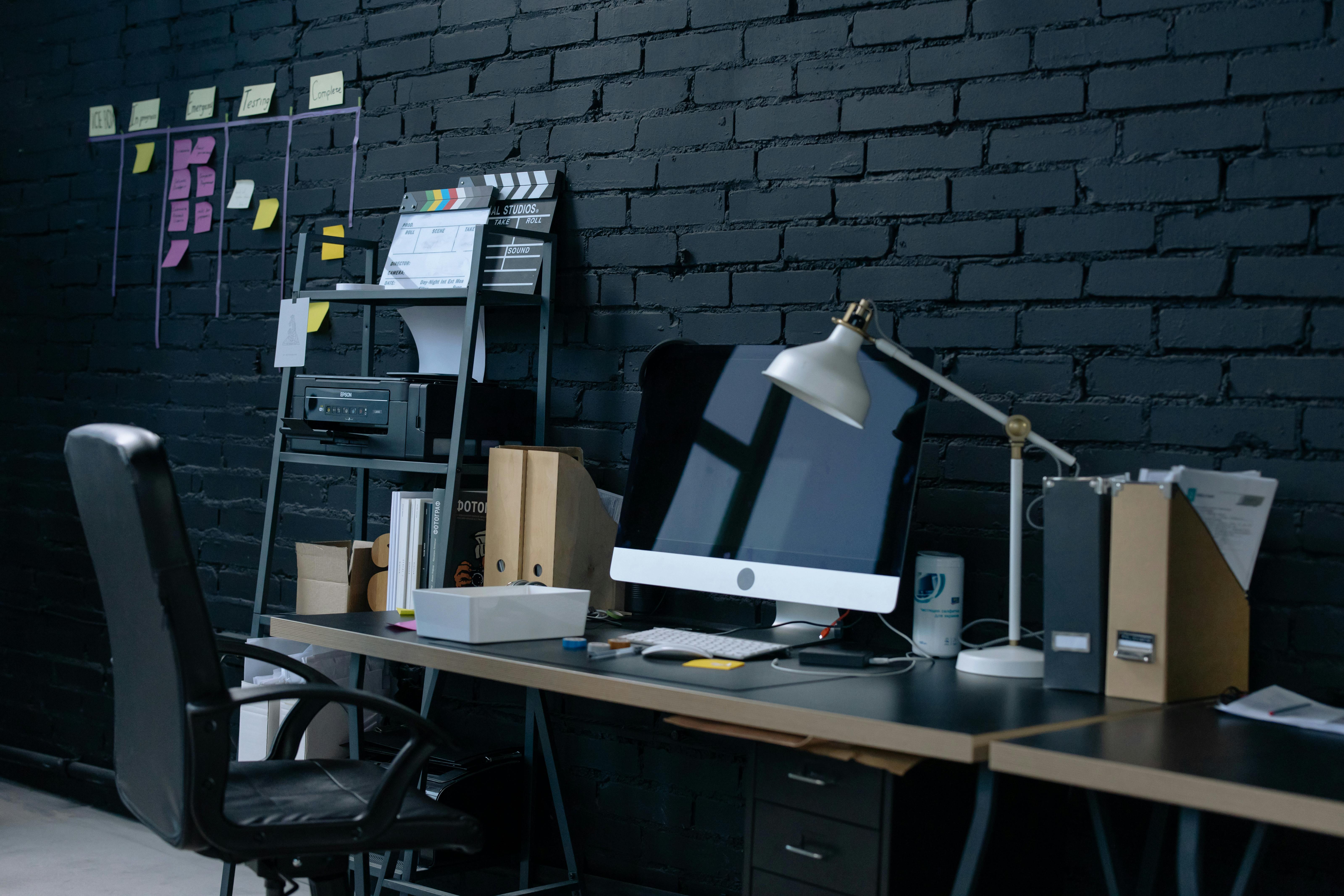 Modern workspace with desktop computer, desk lamp, office chair, and organized files against black brick wall.