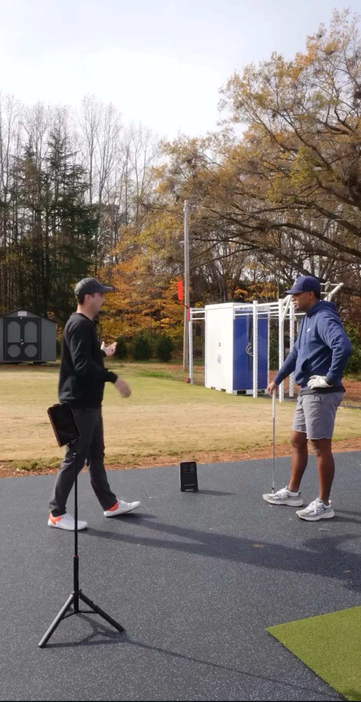 Two men talking on a golf practice area, one holding a putter and wearing a glove, the other gesturing with his hands.