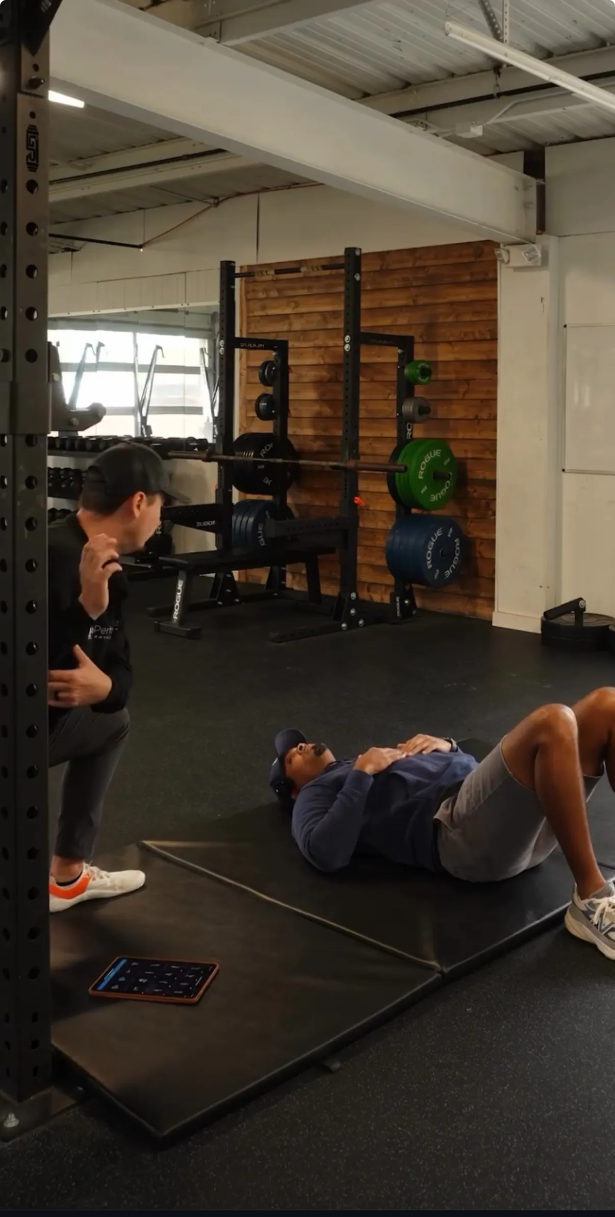 Man lying on a mat in a gym while another person crouches beside him, with weightlifting equipment in the background.
