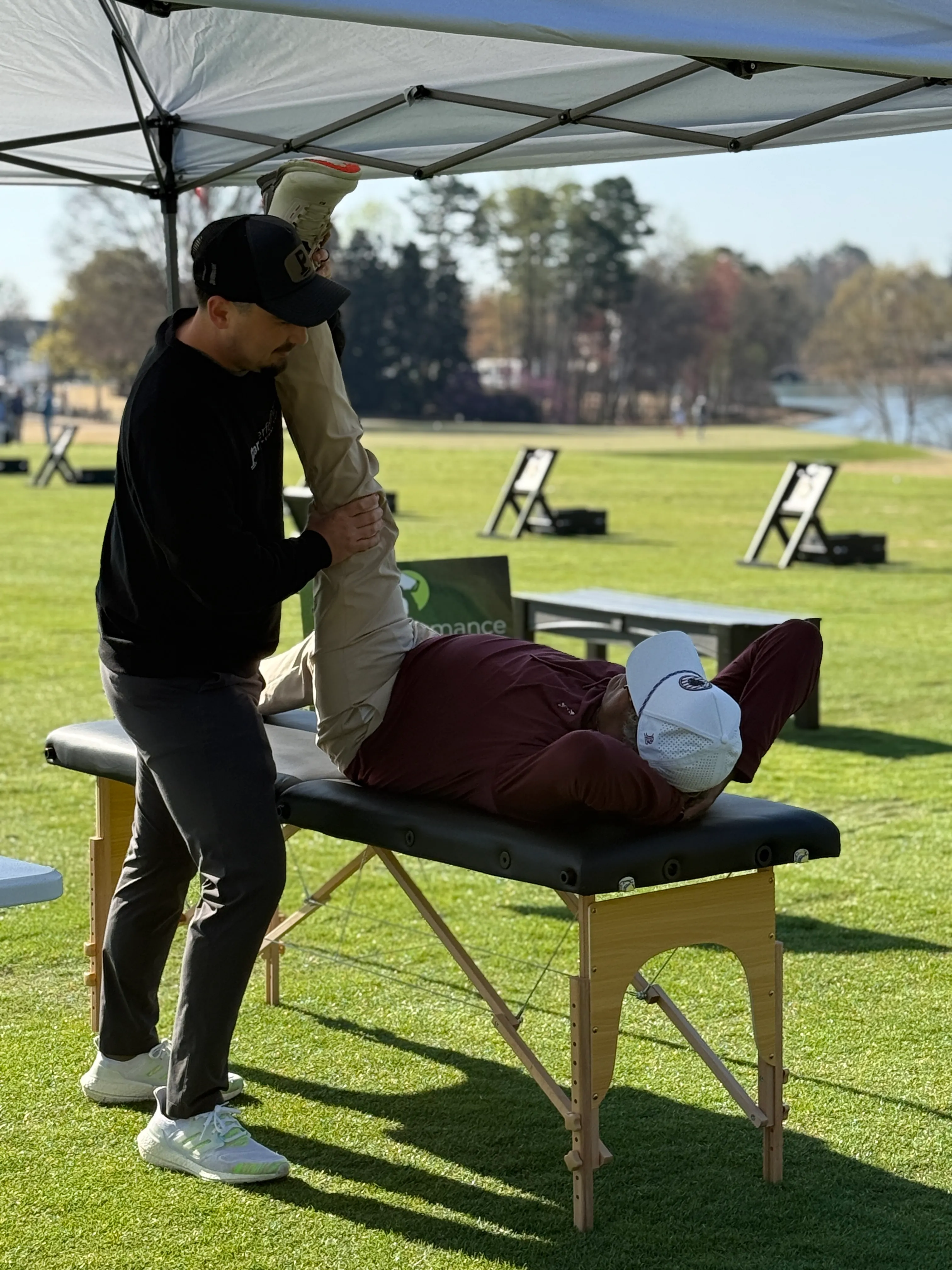 A person lying on a therapy table outdoors while another person stretches their leg under a canopy tent on a grassy field.
